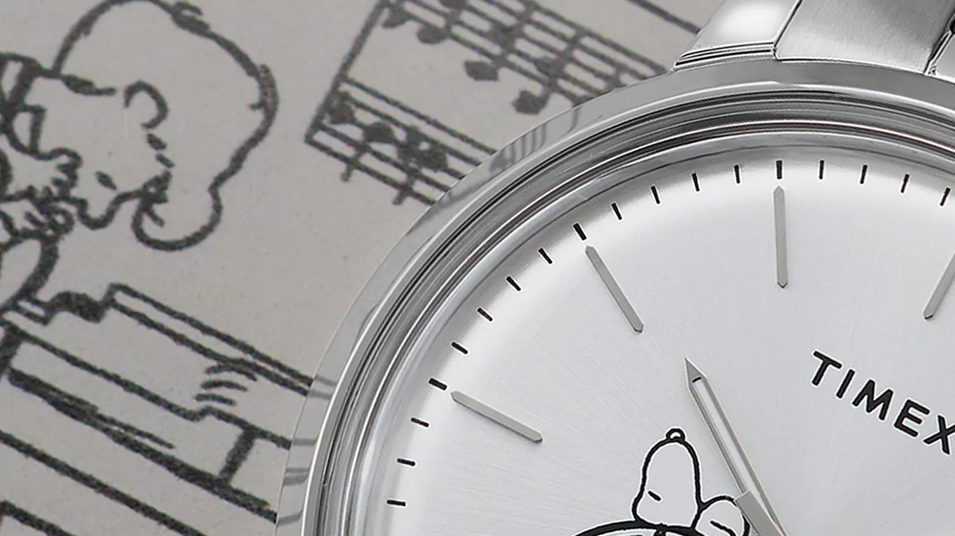 Close-up of a silver Timex wristwatch with a white dial featuring a minimalist design. The watch face includes an illustration of Snoopy from the Peanuts comic strip, positioned near the center. The hour and minute hands are silver, and the watch has a metal band. In the background, there is a blurred black-and-white Peanuts comic strip with musical notes.