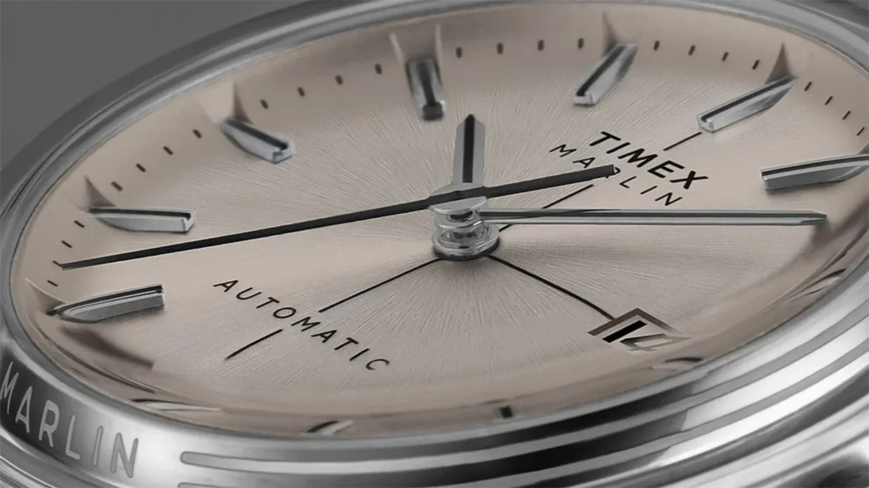 Close-up of a Timex Marlin automatic watch face with a steel dial, steel hour markers, and hands. The watch has a date window at the 3 o'clock position and a black second hand. The bezel is polished silver with "MARLIN" engraved on the side.