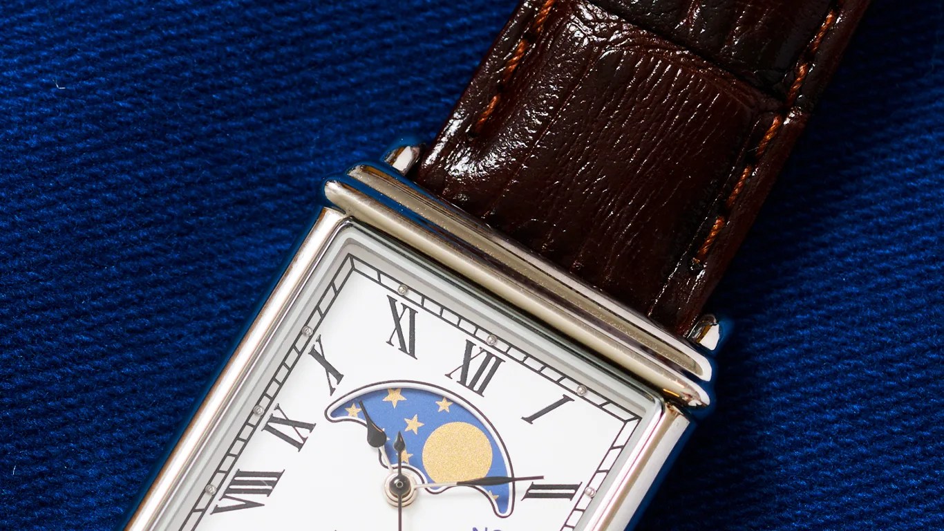 Rectangular timex wristwatch with a white dial featuring black Roman numerals, a moon phase indicator with stars and a golden moon, and a brown leather strap with visible stitching, placed on a textured blue fabric background.