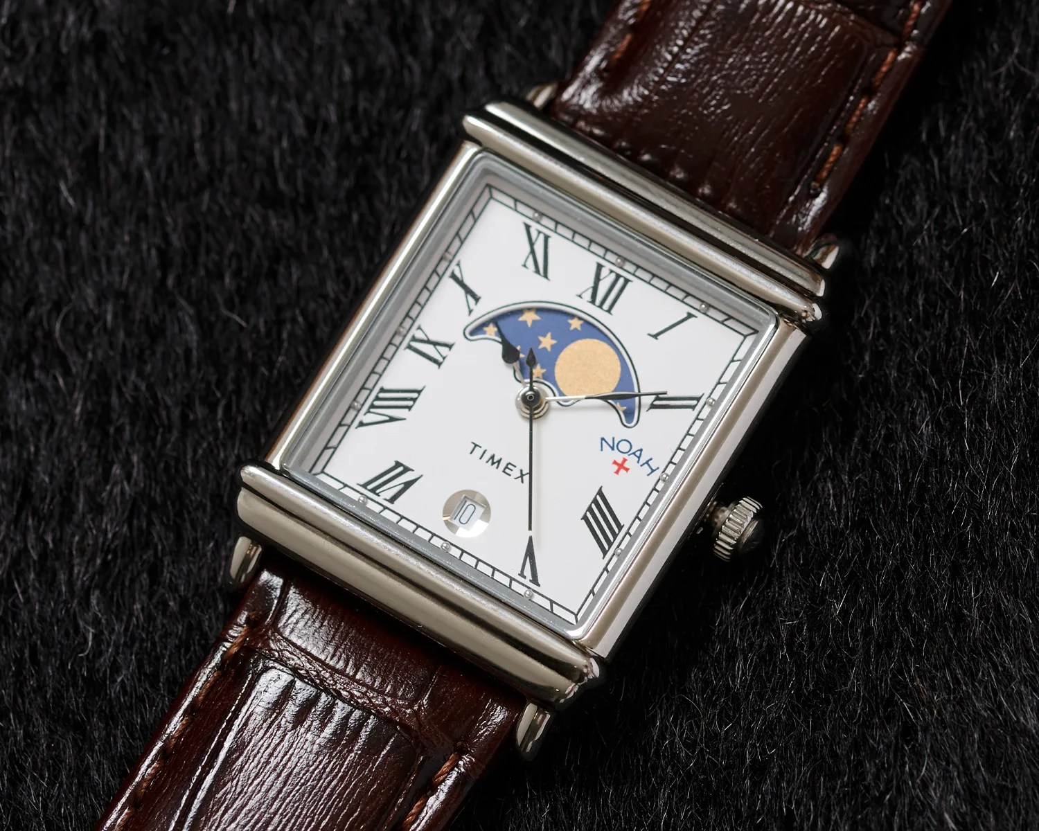 Square silver wristwatch with a white dial featuring black Roman numerals, a moon phase indicator with stars and a gold moon at the top, and a date window at the bottom. The watch has a brown leather strap and the brand names "TIMEX" and "NOAH" are visible on the dial.