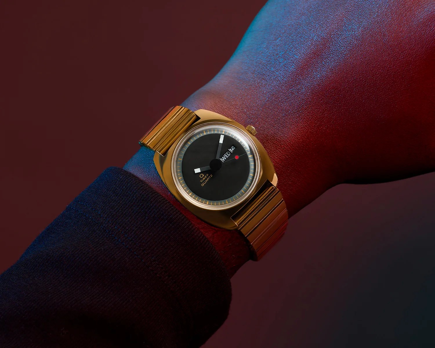 Gold-toned Timex Quartz wristwatch with a black dial, white hour and minute hands, and a red second hand, featuring a day and date display, worn on a wrist with a dark long sleeve.