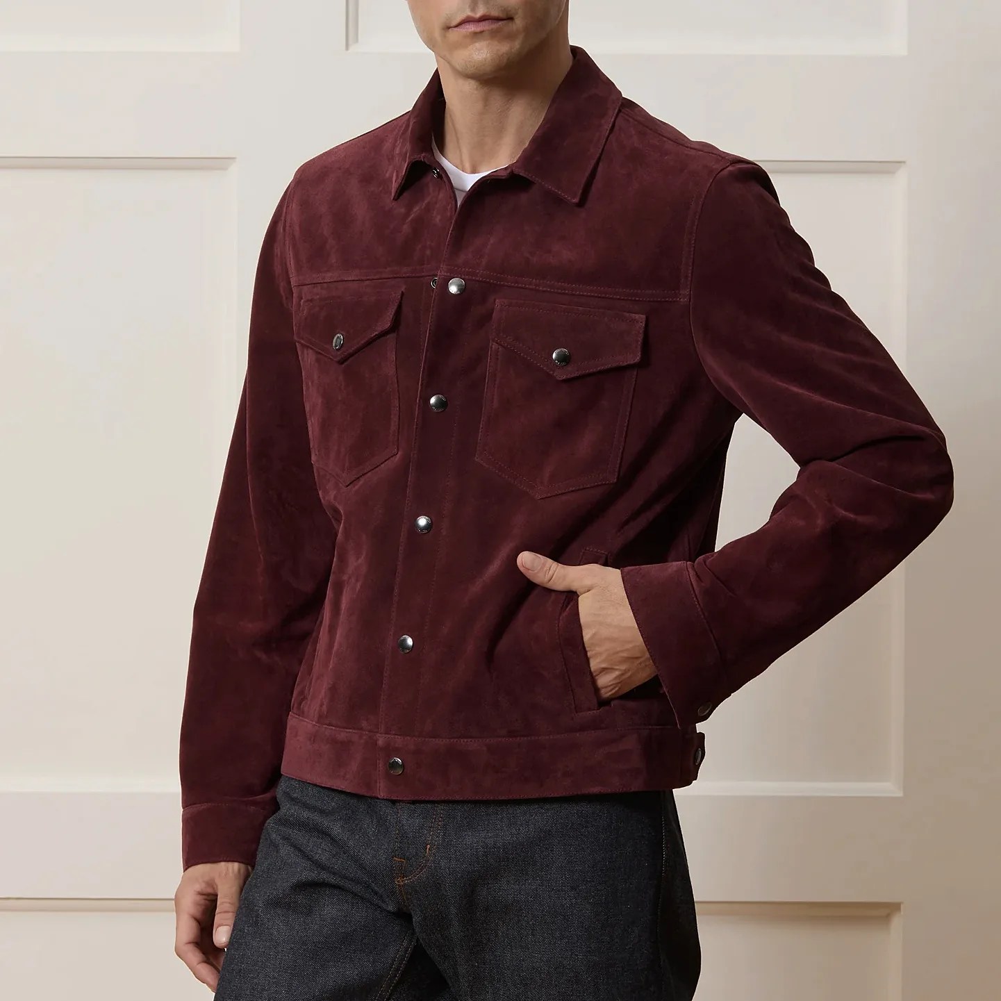A man wearing a dark burgundy suede jacket with a button-up front, two chest flap pockets with buttons, and side pockets. The jacket has a classic collar and buttoned cuffs. He is also wearing dark denim jeans.