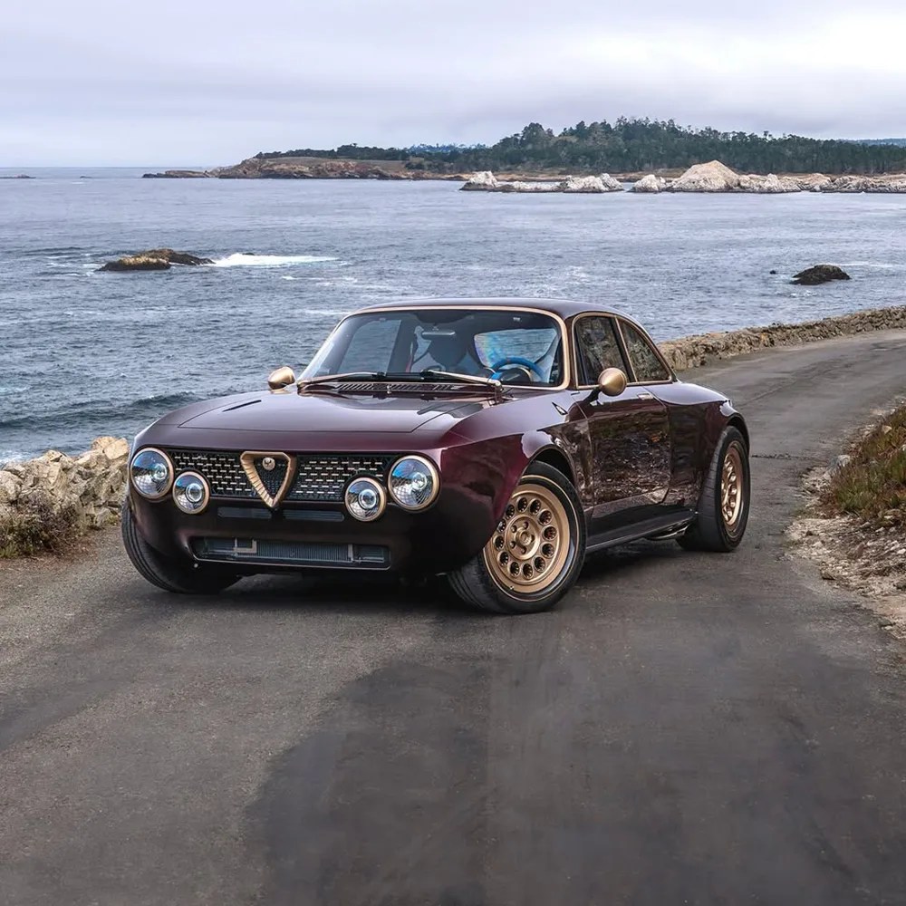Classic dark purple Alfa Romeo sports car with gold accents on the grille, side mirrors, and wheels, parked on a coastal road with the ocean and rocky shoreline in the background.