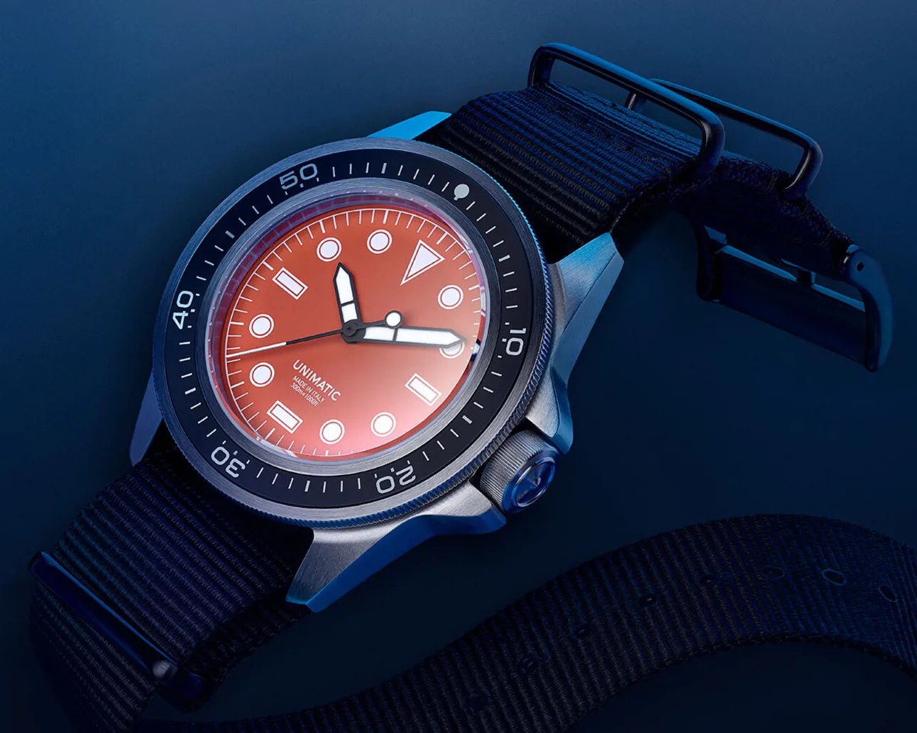 A wristwatch with a bright orange dial featuring white geometric hour markers and thick white hands with black edges. The watch has a black rotating bezel with white minute markings and a stainless steel case. It is paired with a black fabric strap with a black buckle. The dial includes the text "UNIMATIC MADE IN ITALY AUTOMATIC 300m/1000ft.