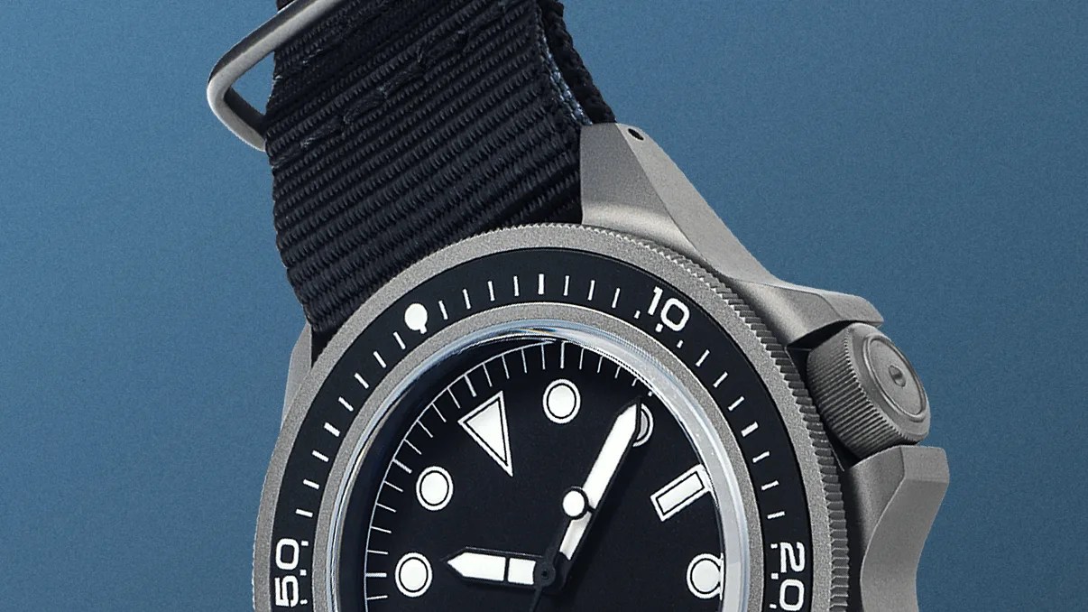 Close-up of a wristwatch with a black dial and white hour markers, featuring a black rotating bezel with white numerals and markings. The watch has a textured crown guard and a black fabric strap with a metal buckle. The hour and minute hands are white with a bold design, and the background is a solid blue color.