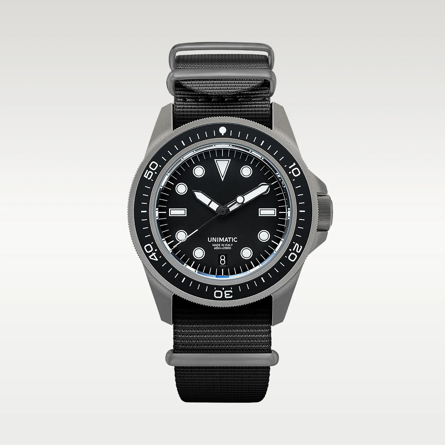 A Unimatic wristwatch with a black dial and white hour markers, featuring a black rotating bezel with white numerals and markings. The watch has a matte grey case and a black nylon strap with metal loops. The dial includes a date window at the 6 o'clock position and the text "UNIMATIC MADE IN ITALY 600m=2000ft." The hour and minute hands are white with black accents, and the second hand is thin and black.