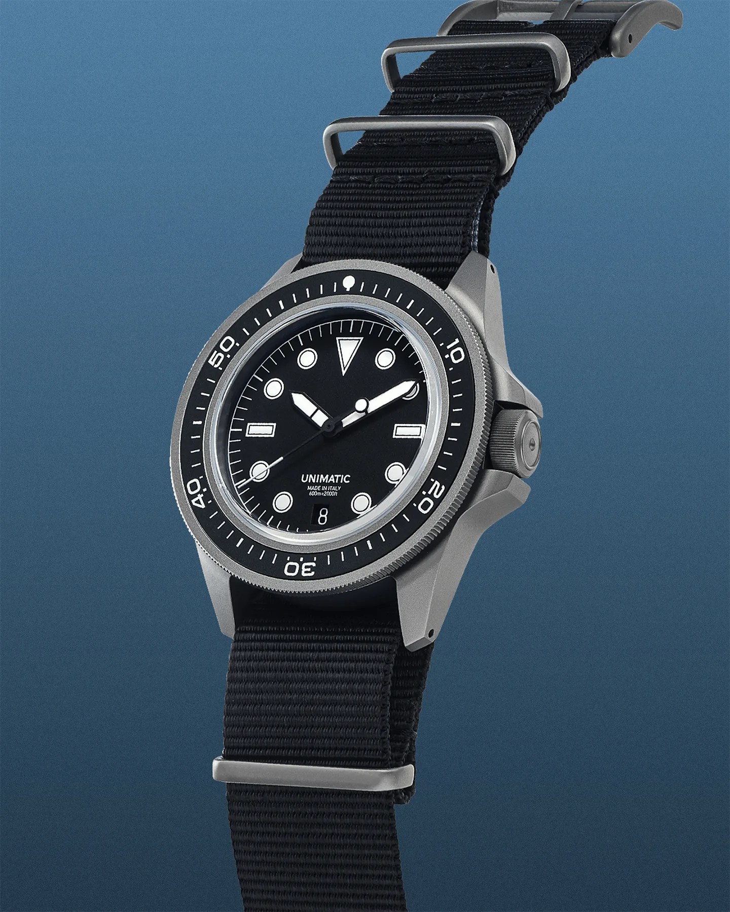 A Unimatic wristwatch with a black dial and white hour markers, featuring a date window at the 6 o'clock position. The watch has a matte silver bezel with minute markings and a black nylon strap with silver hardware. The dial includes the text "UNIMATIC," "MADE IN ITALY," and "600m=2000ft." The watch hands are white with a bold design, and the second hand has a circular tip.