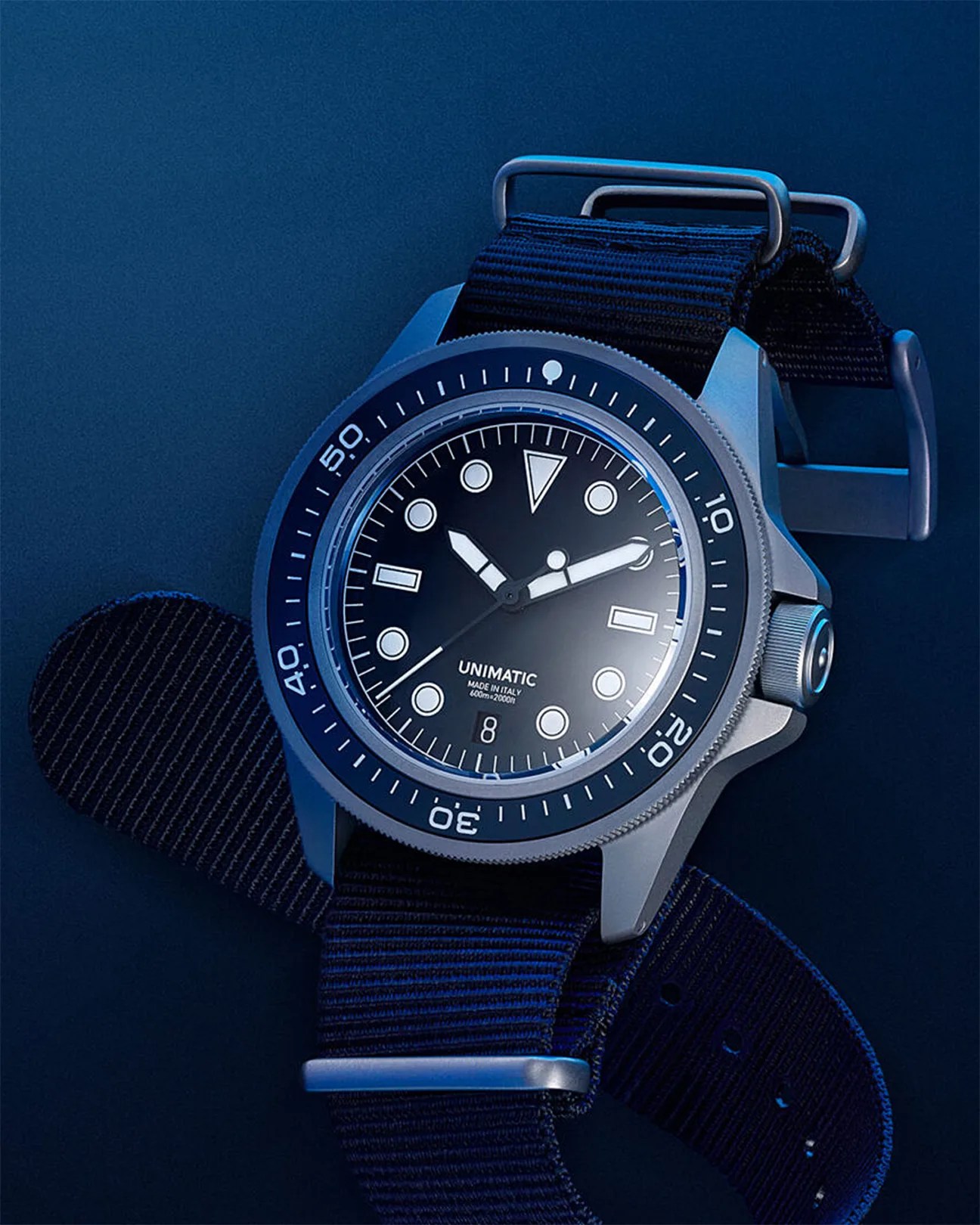 A close-up of a Unimatic wristwatch with a black dial and white hour markers, featuring a rotating bezel with minute markings. The watch has a black fabric strap with metal loops and a date window at the 6 o'clock position. The watch case is metallic with a textured crown on the right side. The dial indicates "UNIMATIC," "MADE IN ITALY," and "600m=2000ft.
