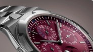 Close-up of a luxury wristwatch with a brushed stainless steel case and bracelet. The watch face is a deep burgundy color with multiple subdials displaying months, days of the week, and dates. The hour markers are silver and rectangular, with white minute markers and blue numerals on the outer edge. The watch hands are silver with a sleek, modern design. The dial also features a moon phase indicator at the bottom.