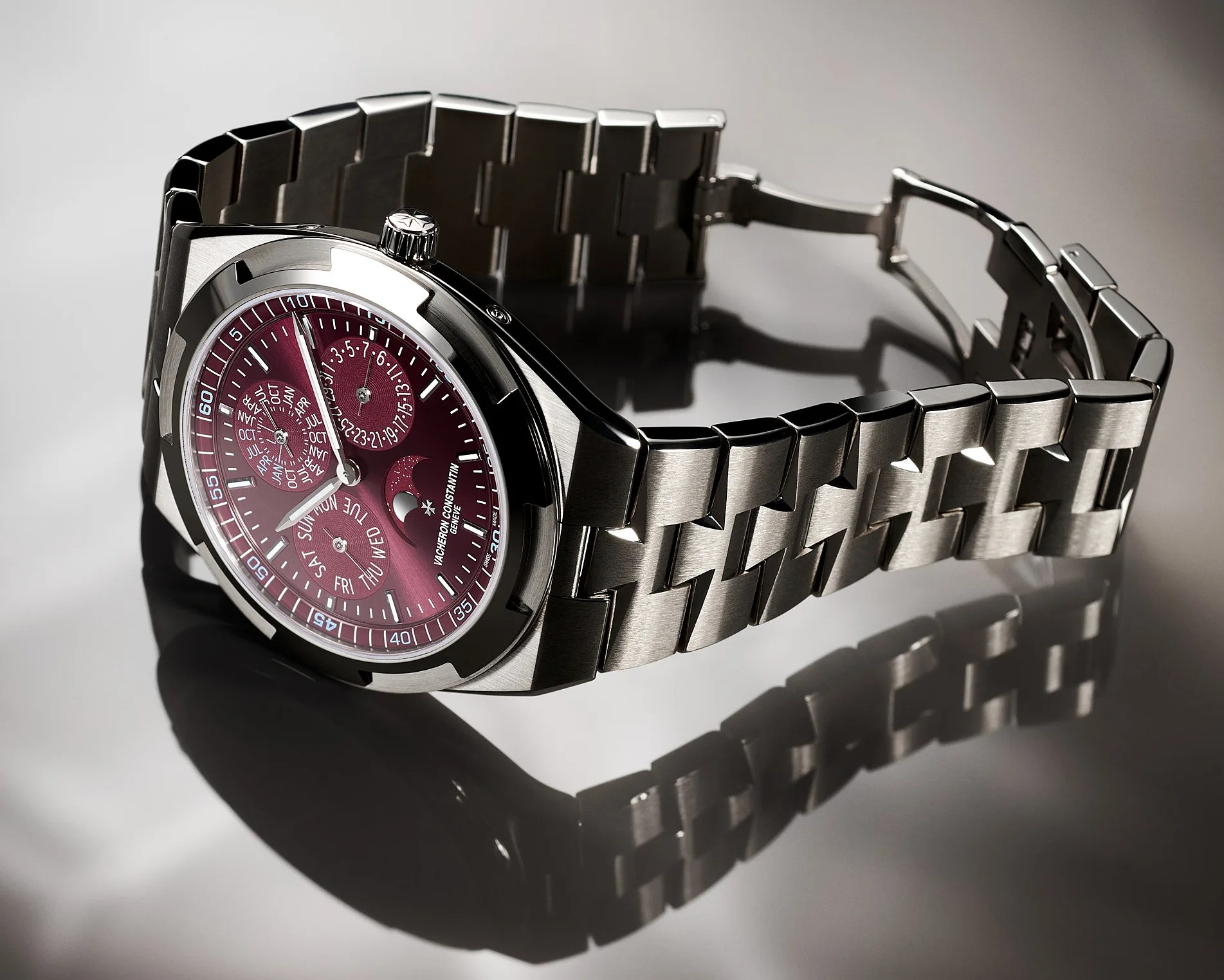 Stainless steel wristwatch with a burgundy dial featuring multiple subdials for day, date, month, and moon phase, and a linked metal bracelet. The watch has a polished and brushed finish with a prominent crown and a reflective surface beneath.