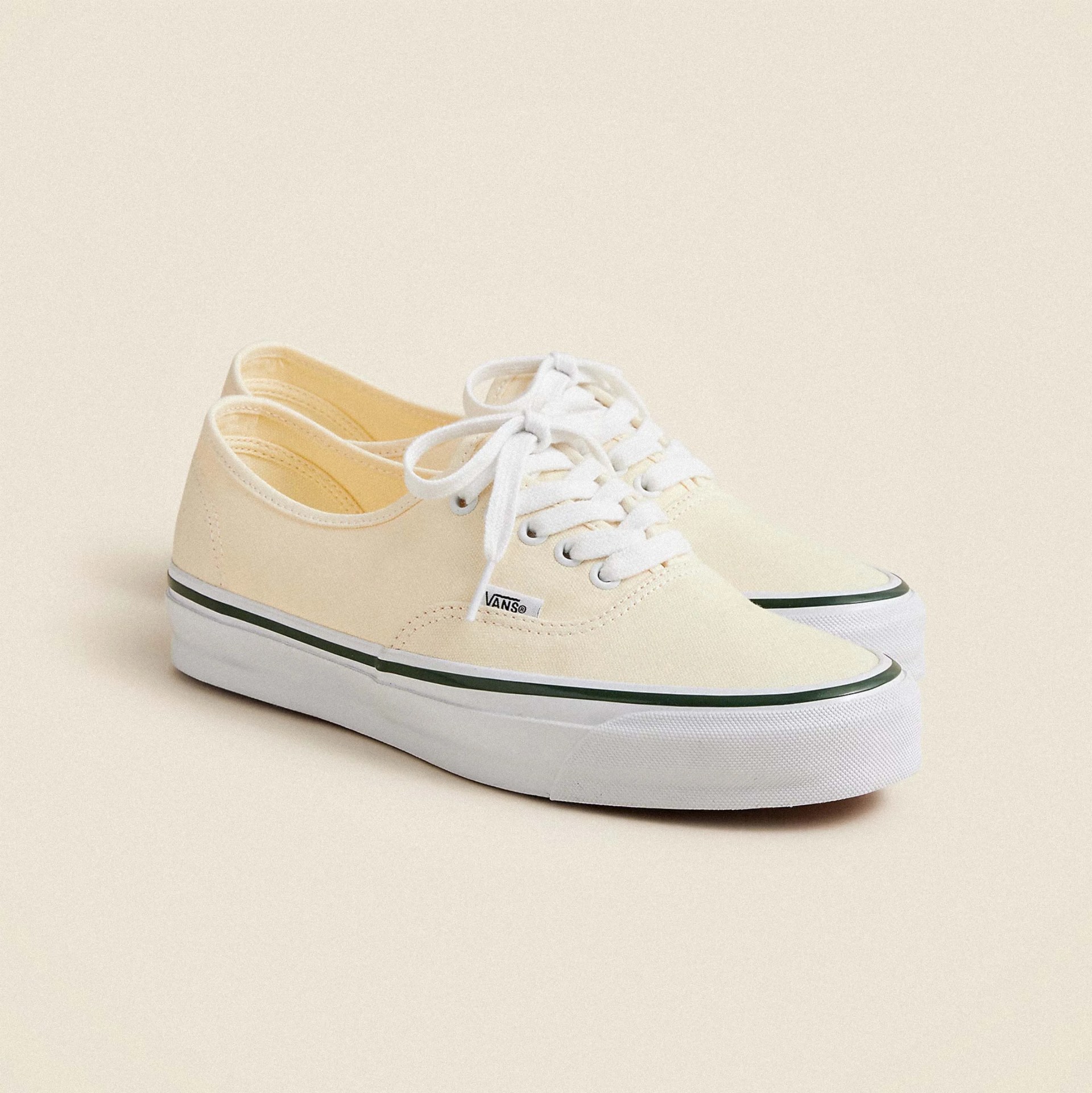 A pair of off-white Vans sneakers with white laces and a white rubber sole featuring a thin black stripe near the top edge. The shoes have a low-top design and a small Vans logo tag on the side.