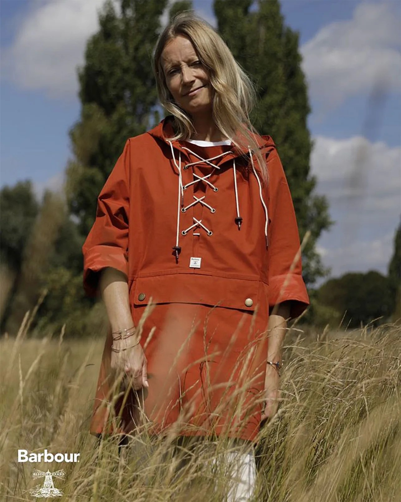 A woman stands in a field of tall grass wearing a rust-colored Barbour jacket with a lace-up front and three-quarter sleeves. The jacket features a front flap pocket with button details. The background includes tall green trees and a partly cloudy sky.