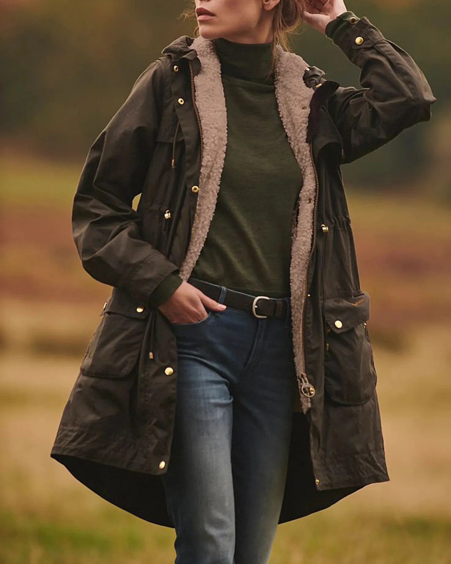 Dark olive green long parka jacket with a beige fleece lining, worn over a dark green turtleneck sweater and blue jeans with a black belt. The jacket features gold buttons and large front pockets. The background is an outdoor setting with blurred natural scenery.