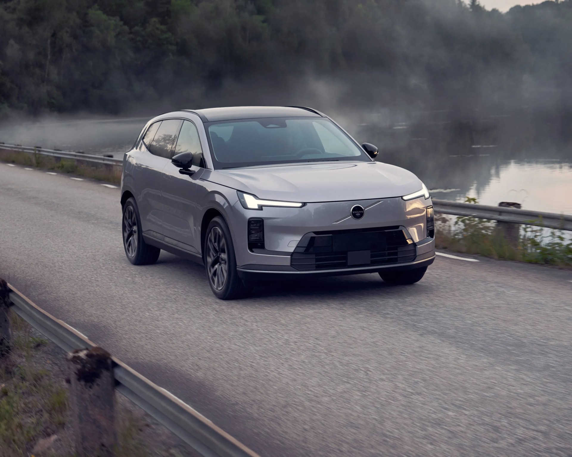 Silver Volvo electric SUV driving on a paved road beside a body of water with mist and trees in the background. The vehicle features sleek, modern headlights and a black roof.