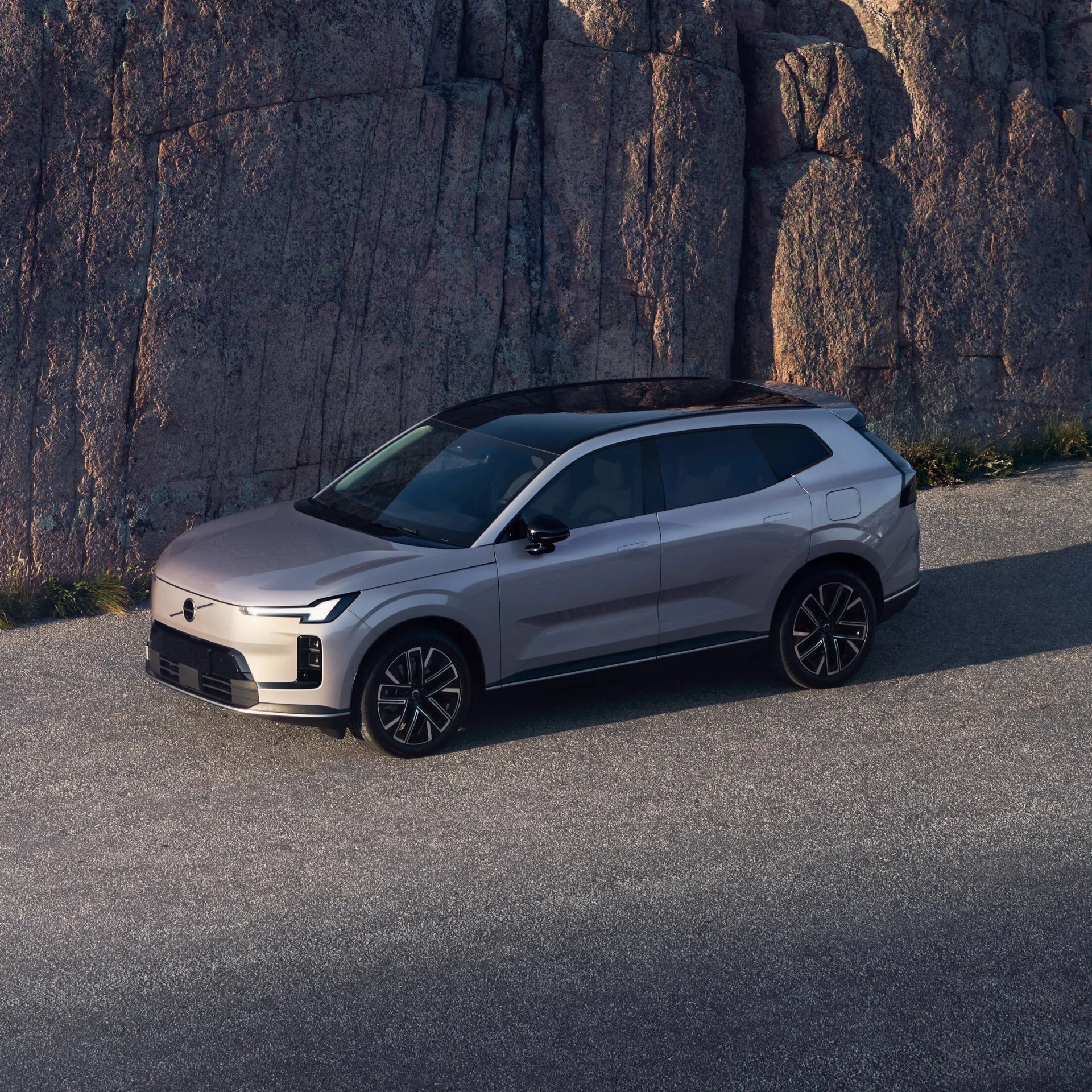 A silver modern SUV with a black roof is parked on a paved surface in front of a large rock wall. The vehicle features sleek, angular headlights and black alloy wheels. The setting appears to be outdoors in natural light.
