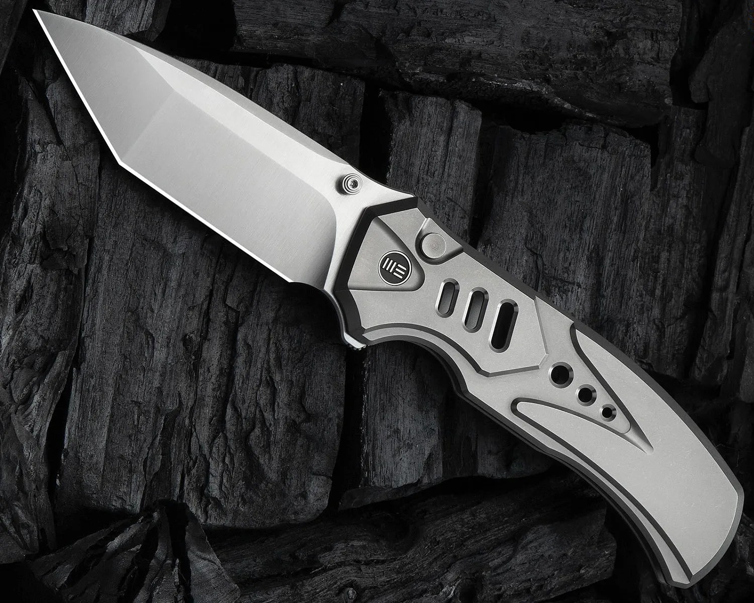 A folding knife with a sleek, modern design featuring a silver blade and a matching silver handle. The blade has a sharp, angular shape with a smooth finish. The handle has geometric cutouts and a circular button near the blade's base, with a small logo consisting of three horizontal lines inside a circle. The knife is placed on a dark, textured wooden surface.