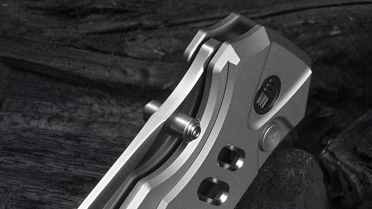 Close-up of a metallic folding knife handle with a clip and a pivot screw, showing a sleek, brushed finish and precise machining details. The background is dark and textured.