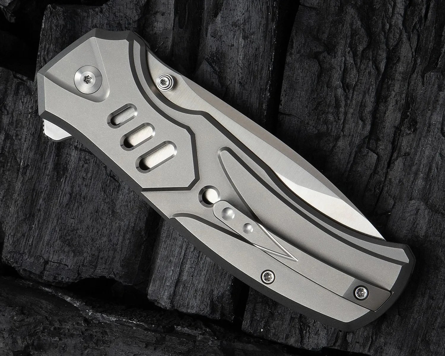A closed folding knife with a metallic silver handle featuring geometric cutouts and a clip, resting on a textured black surface. The blade is partially visible, tucked inside the handle.
