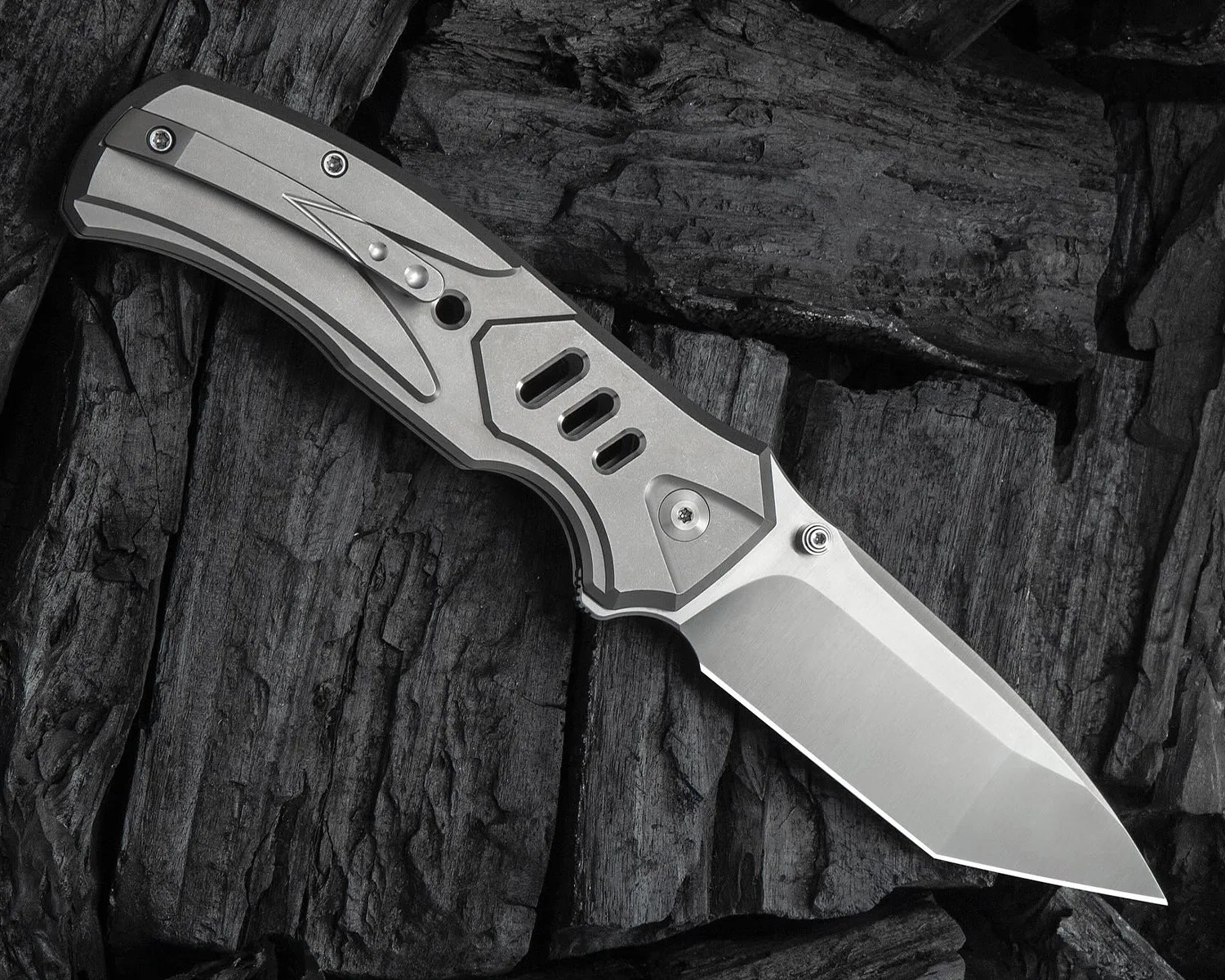 Folding knife with a metallic handle featuring geometric cutouts and a clip on one side. The blade is partially open, has a sharp angular tip, and a smooth, polished finish. The background consists of dark, textured wood pieces.