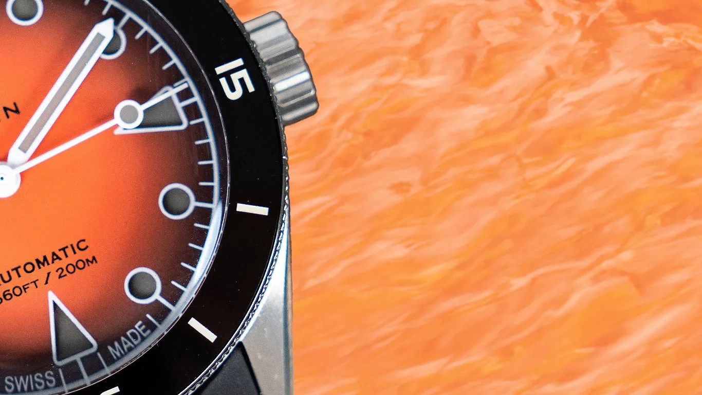 Close-up of a wren watch face with an orange gradient dial, black bezel, and white markings. The dial shows "AUTOMATIC 660FT / 200M" and "SWISS MADE." The hour and minute hands are white with a bold design, and the second hand is thin and white. The watch crown is visible on the right side. The background is an orange textured surface.