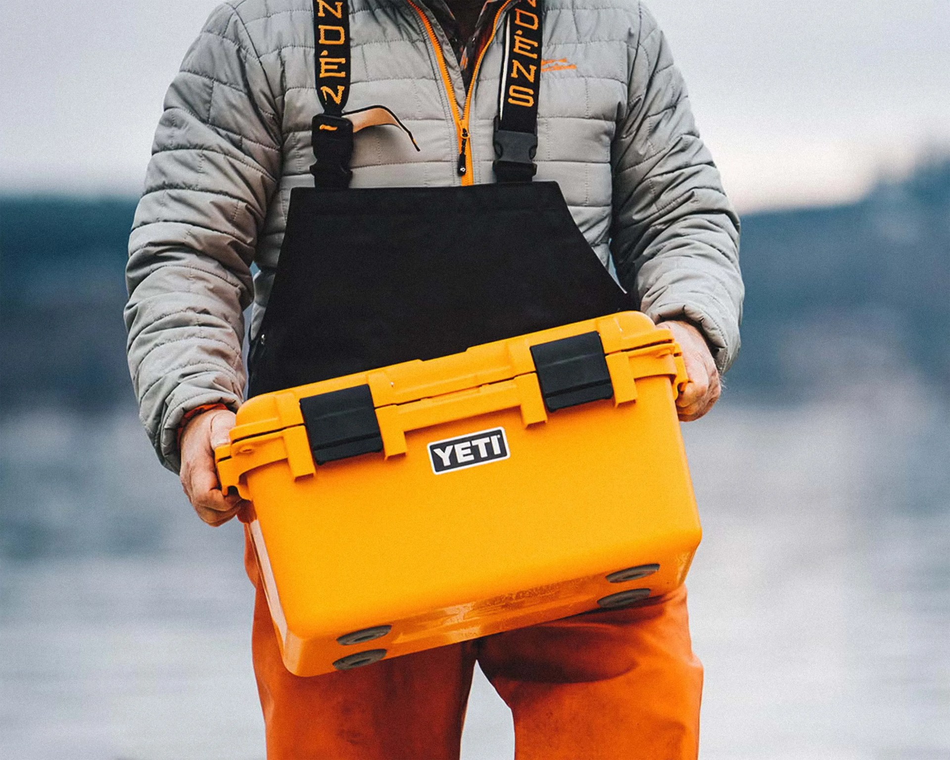 A person wearing a gray quilted jacket, black bib overalls and orange pants is holding a bright yellow YETI cooler with black latches. The background is blurred, showing an outdoor setting near water.