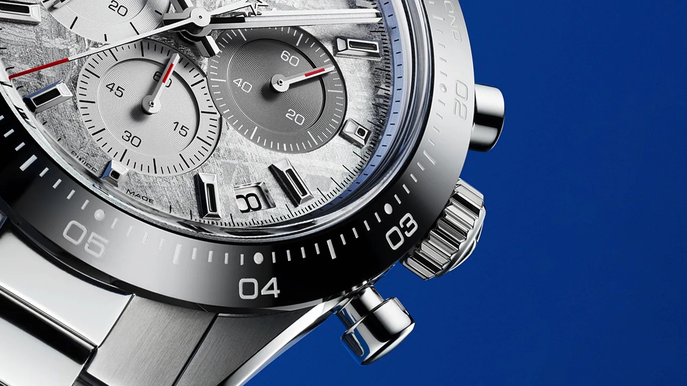 Close-up of a luxury zenith chronograph watch with a textured silver dial, three subdials, a date window, and a black bezel with white markings. The watch has a stainless steel case and bracelet, with a blue background.