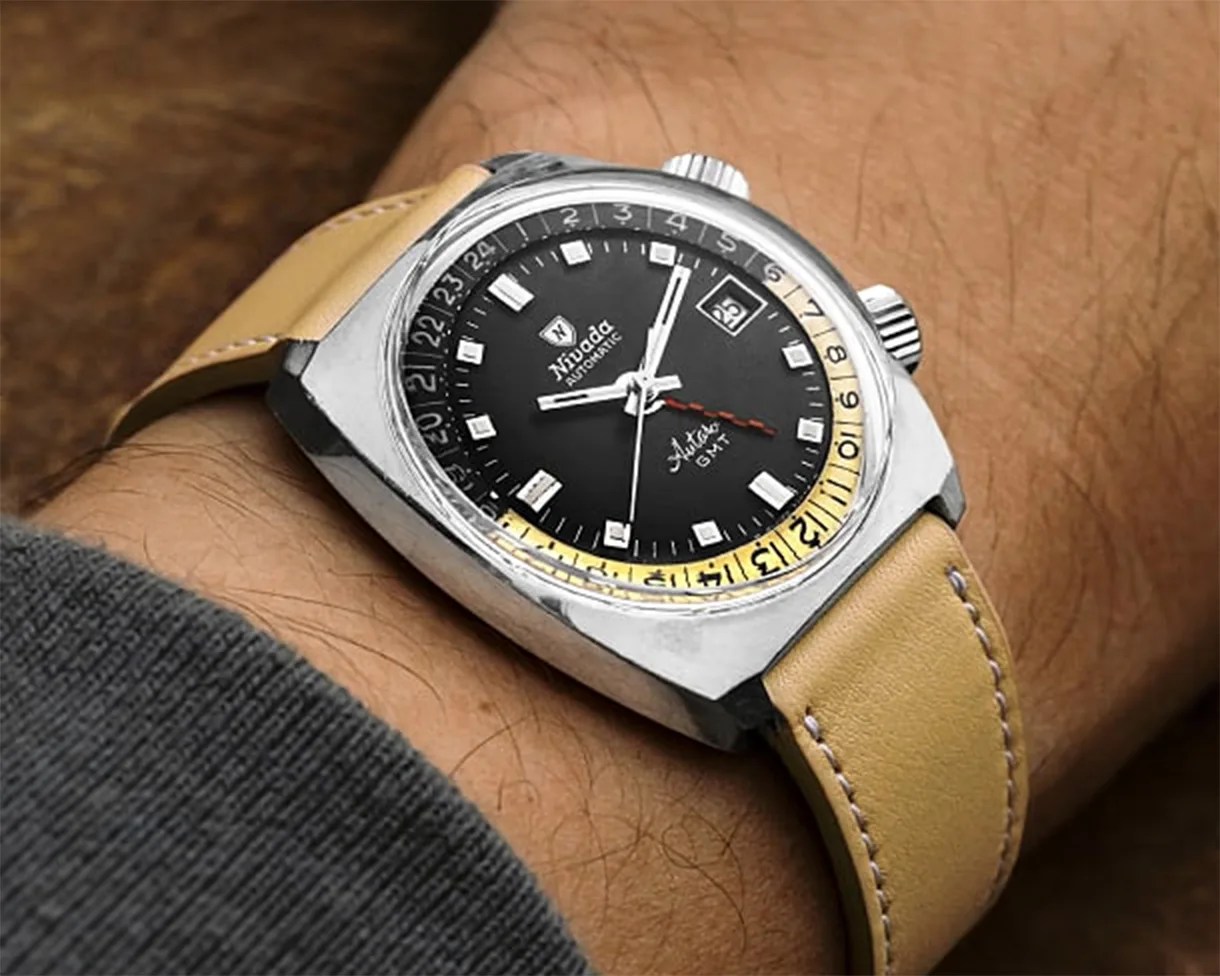 Silver wristwatch with a black dial and beige leather strap, featuring rectangular hour markers, a date window at 3 o'clock, and a yellow 24-hour GMT scale around the edge. The watch has two crowns and a red GMT hand.