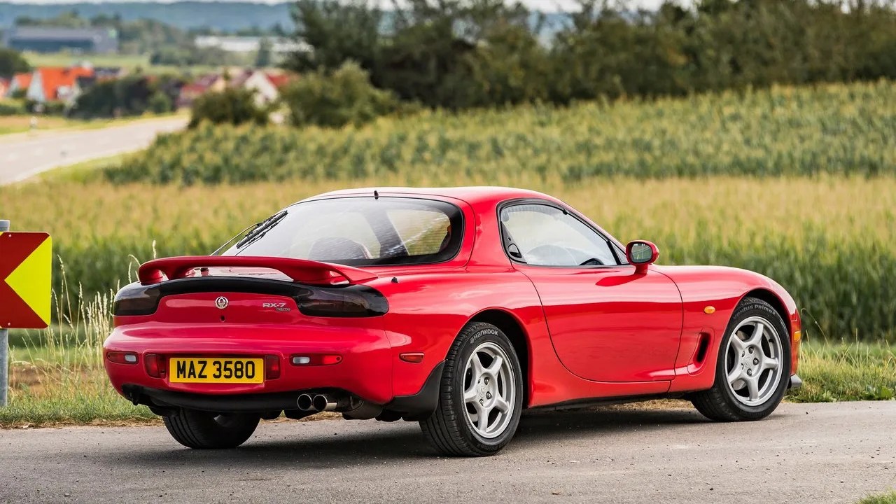 Red Mazda RX-7 sports car parked on a rural road with green fields and trees in the background, featuring a rear spoiler, dual exhaust pipes, and silver alloy wheels.