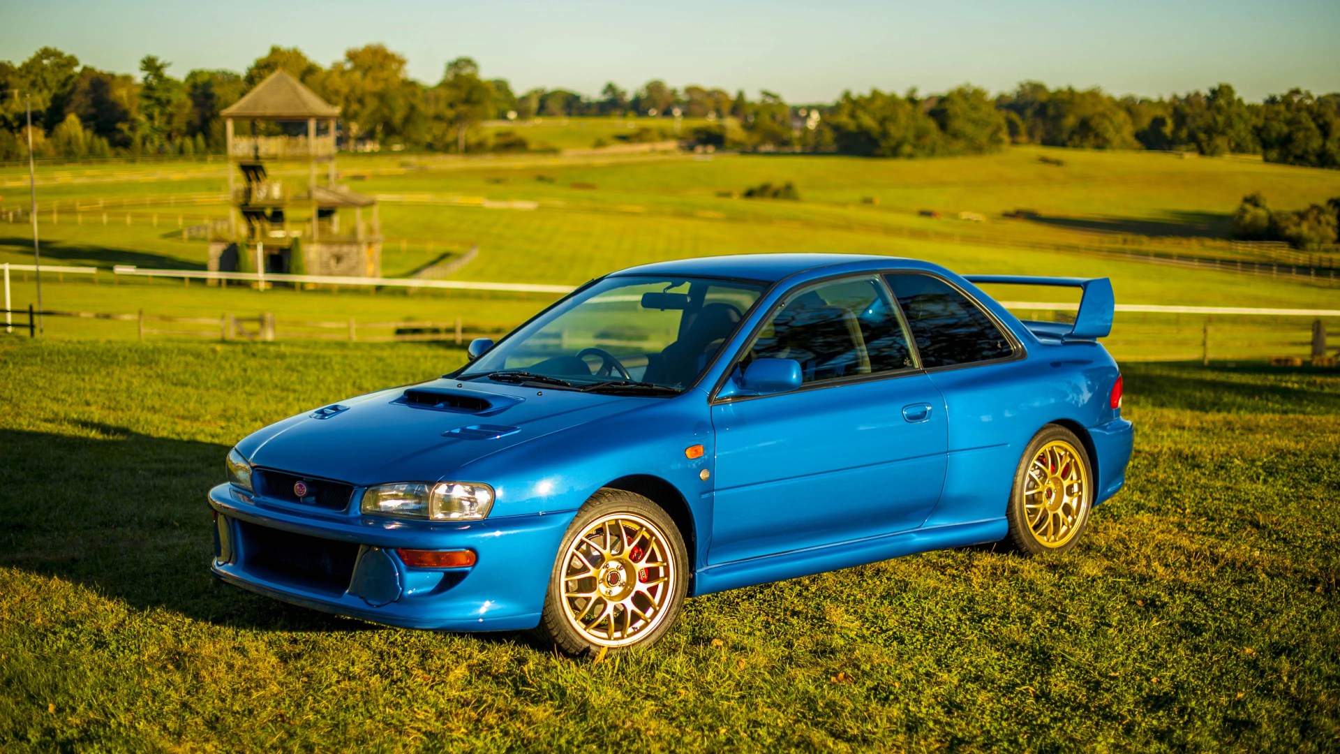 Blue Subaru Impreza WRX with gold alloy wheels parked on grass in a sunny, rural setting with a wooden structure and trees in the background.