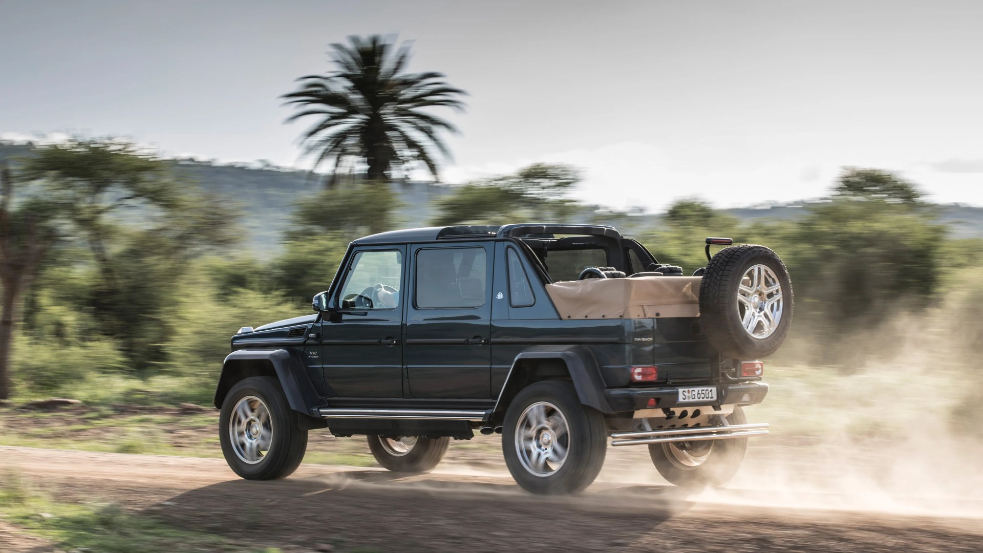 Dark green Mercedes-Benz G-Class 4x4 SUV driving on a dusty dirt road with a beige soft top covering the rear cargo area and a spare tire mounted on the back. The vehicle has a V12 biturbo badge on the front fender and is surrounded by a natural landscape with trees and a palm tree in the background.
