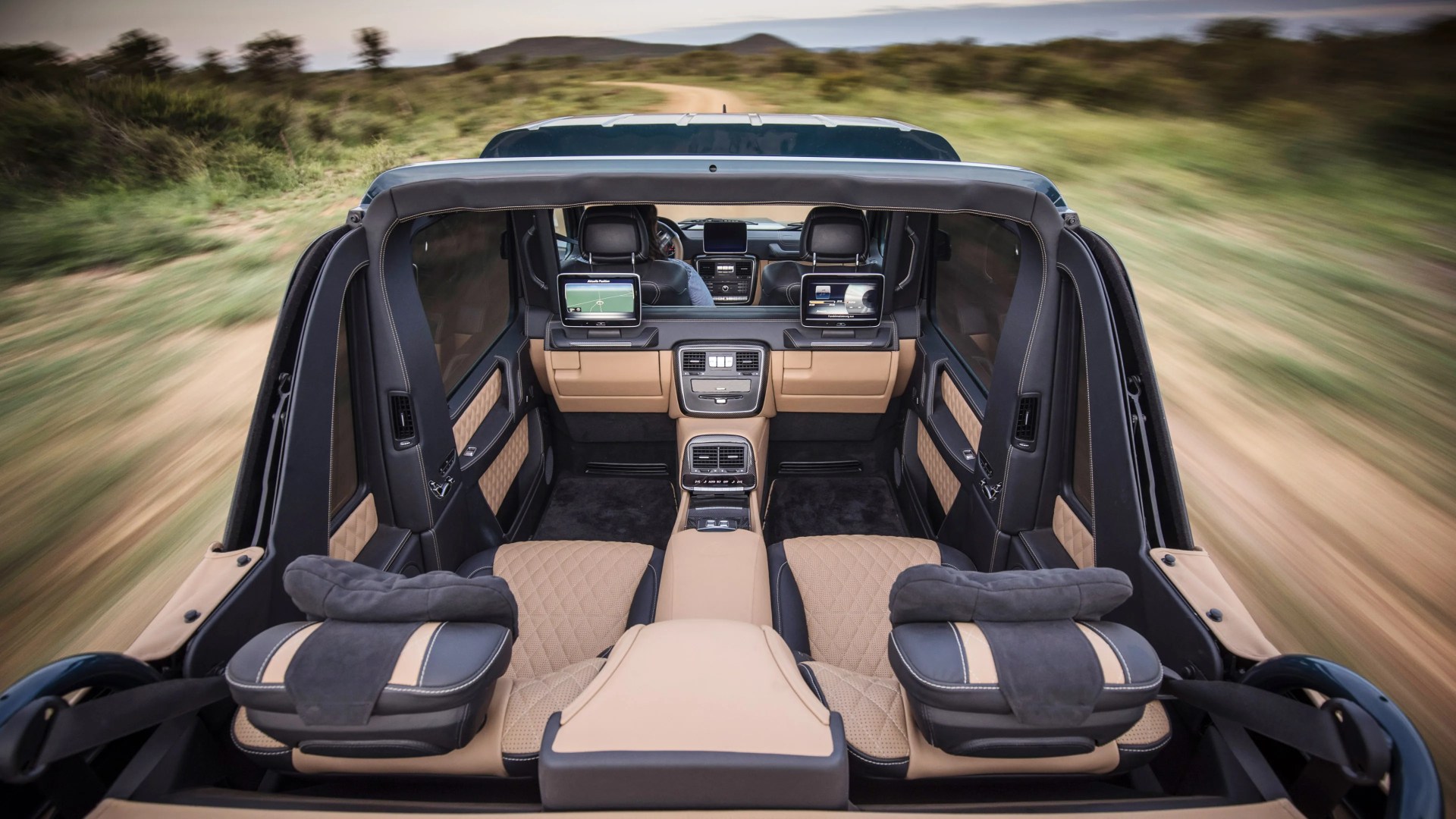 Rear interior view of a luxury vehicle with beige and black quilted leather seats, two headrest-mounted screens, and a central console featuring air vents and controls, driving on a dirt road surrounded by greenery.