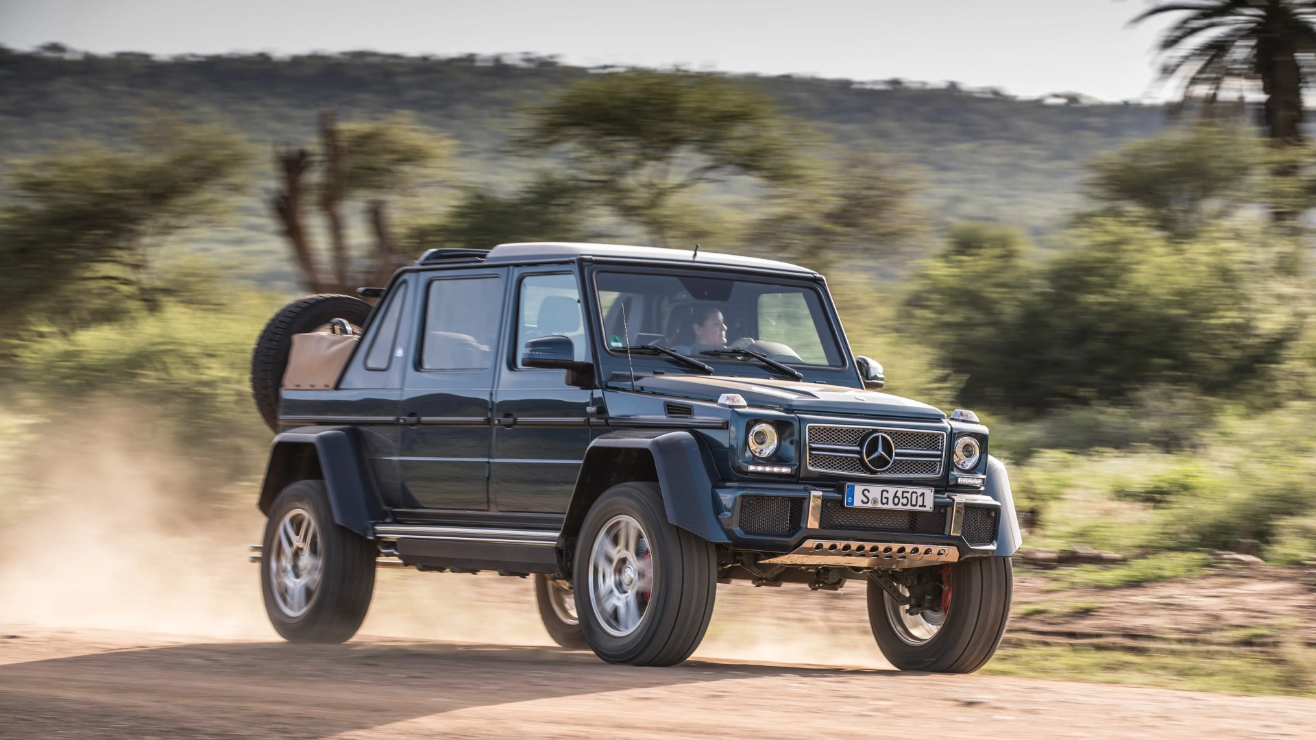 A dark-colored Mercedes-Benz G-Class SUV driving on a dirt road, kicking up dust. The vehicle has a spare tire mounted on the rear and a tan cover over the rear cargo area. The background features green trees and hills under a clear sky.