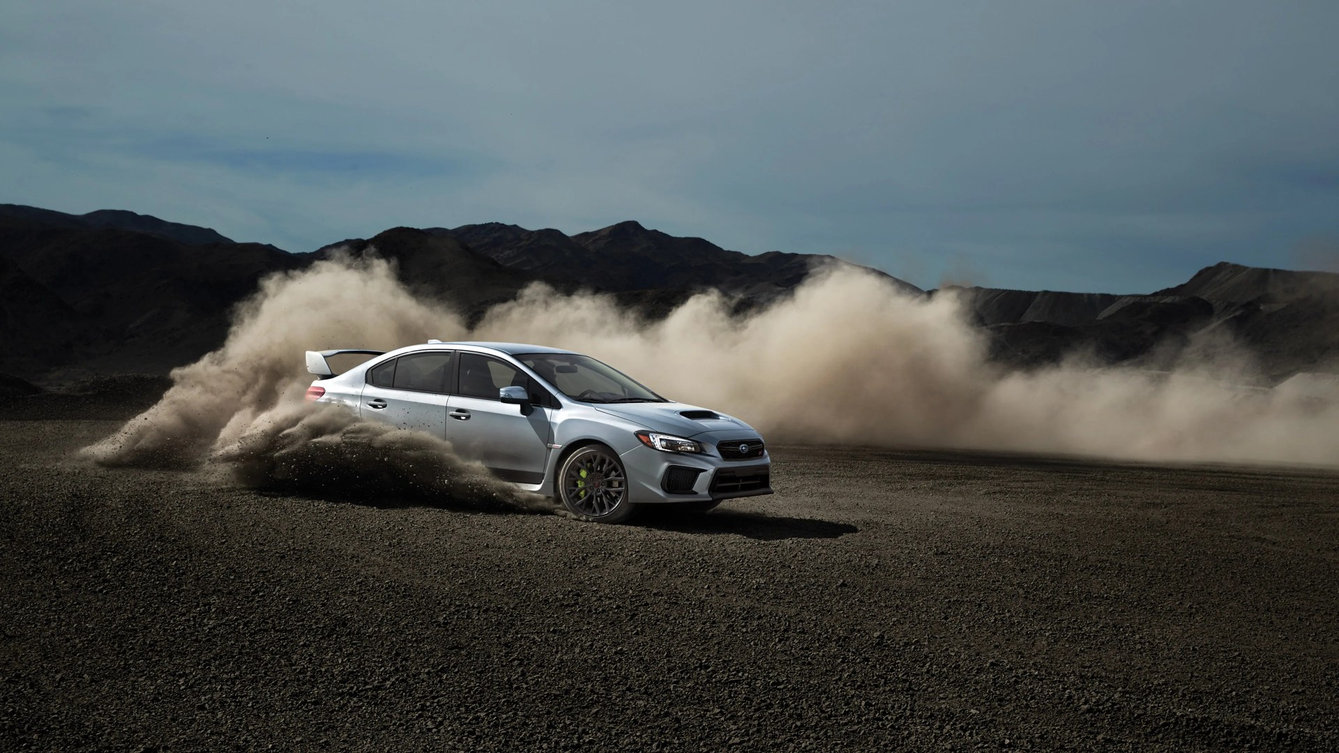 Silver Subaru WRX sedan drifting on a gravel surface, kicking up a large cloud of dust with mountains in the background under a clear sky.