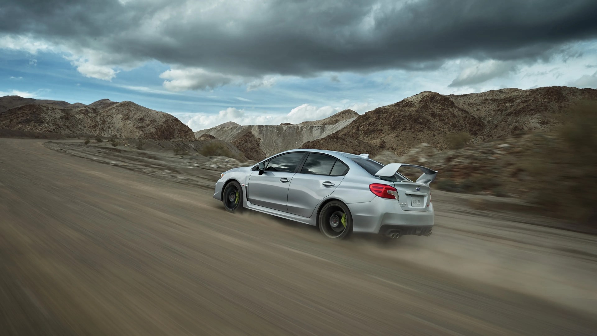 Silver Subaru WRX sedan with a large rear spoiler driving at speed on a dusty dirt road in a mountainous desert area under a cloudy sky.