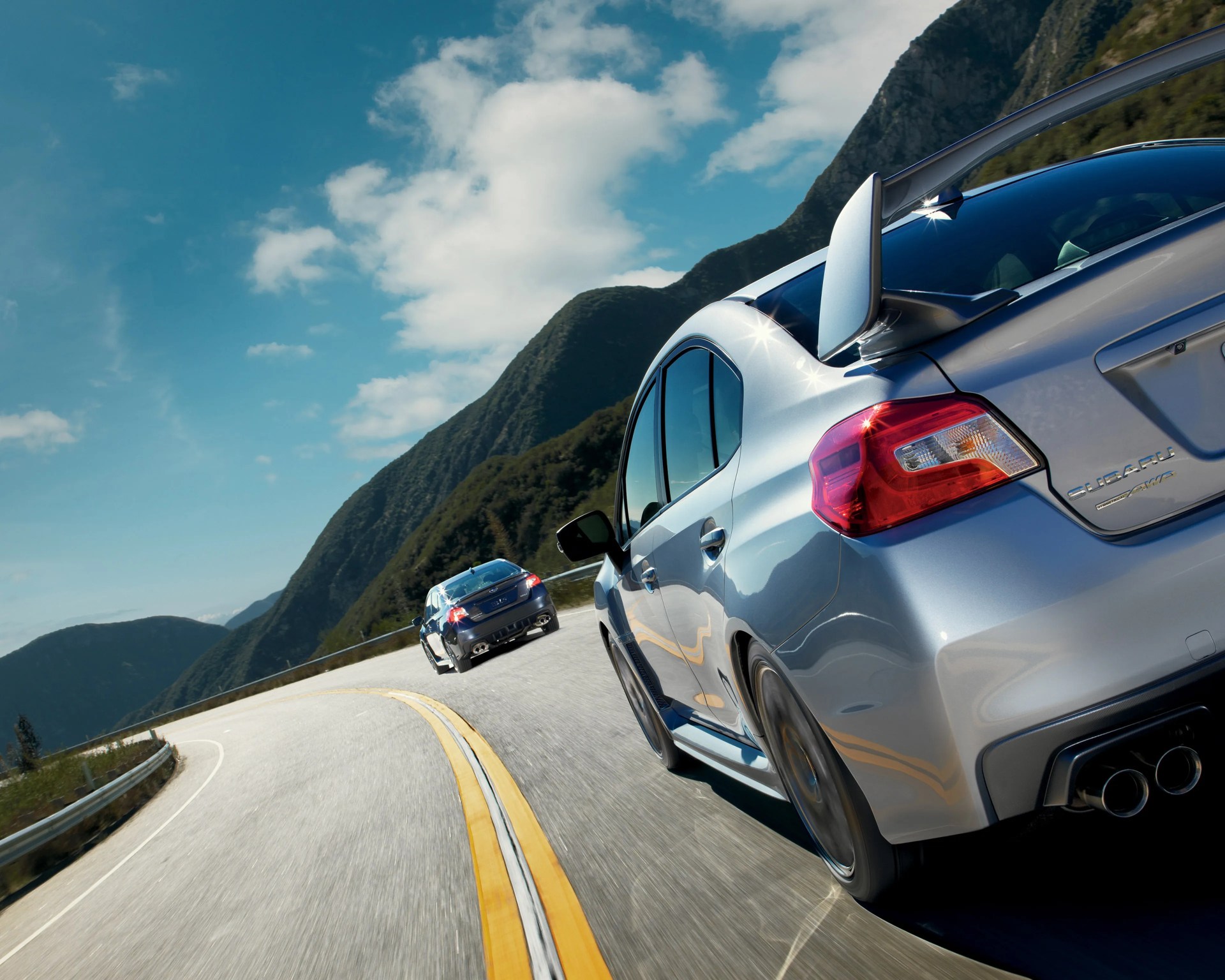 Two silver Subaru WRX STI cars driving on a winding mountain road under a partly cloudy blue sky, with green forested hills in the background. The focus is on the rear side of the closest car, showing its spoiler, taillight, and dual exhaust.