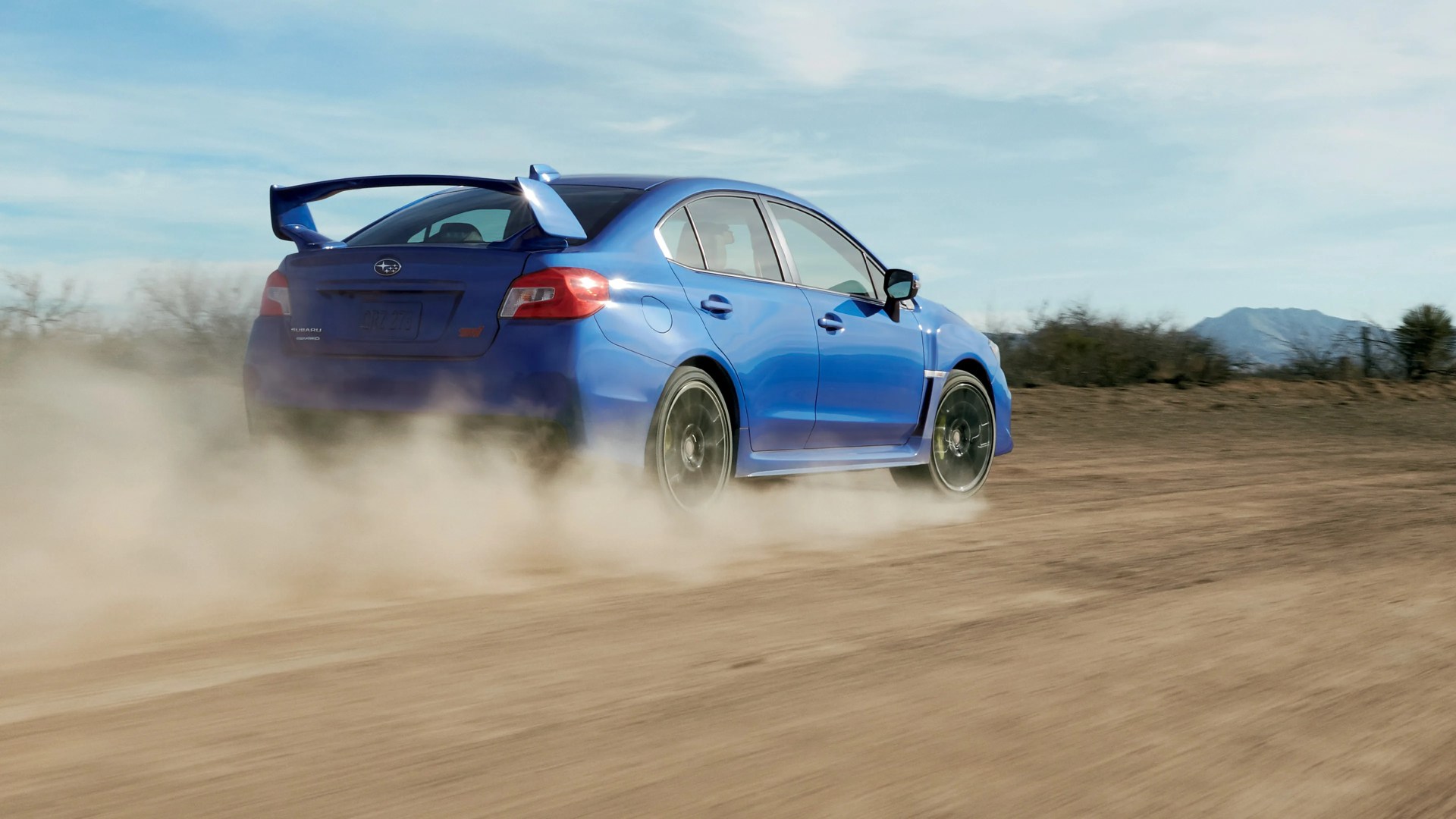 Blue Subaru WRX STI sedan driving on a dirt road, kicking up dust behind it, with a clear sky and distant mountains in the background.
