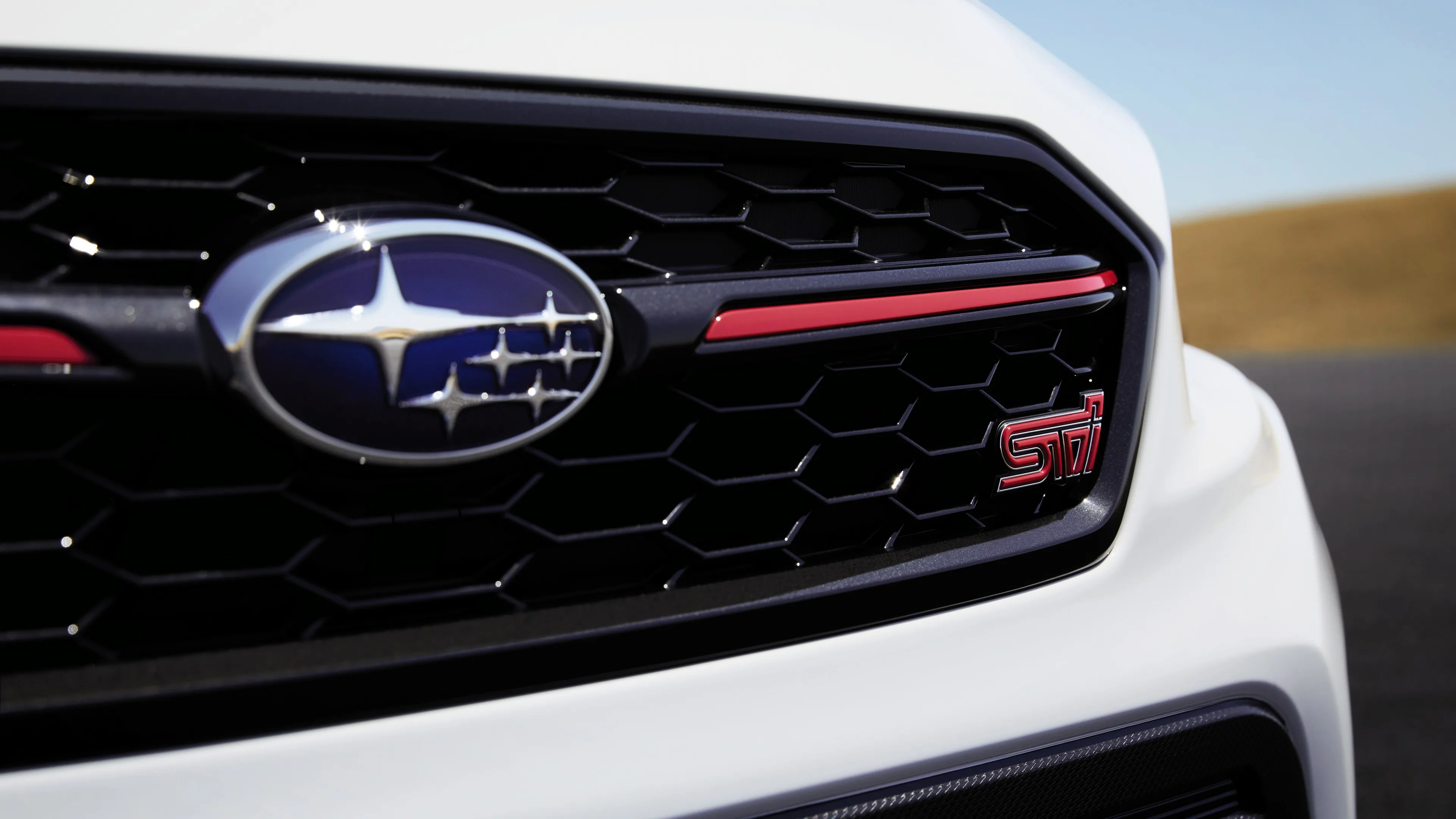 Close-up of a white Subaru car front grille featuring the Subaru emblem on the left and a red "STI" badge on the right, with a honeycomb pattern and a red accent line above the badge.
