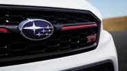 Close-up of a white Subaru car front grille featuring the Subaru emblem on the left and a red "STI" badge on the right, with a honeycomb pattern and a red accent line above the badge.