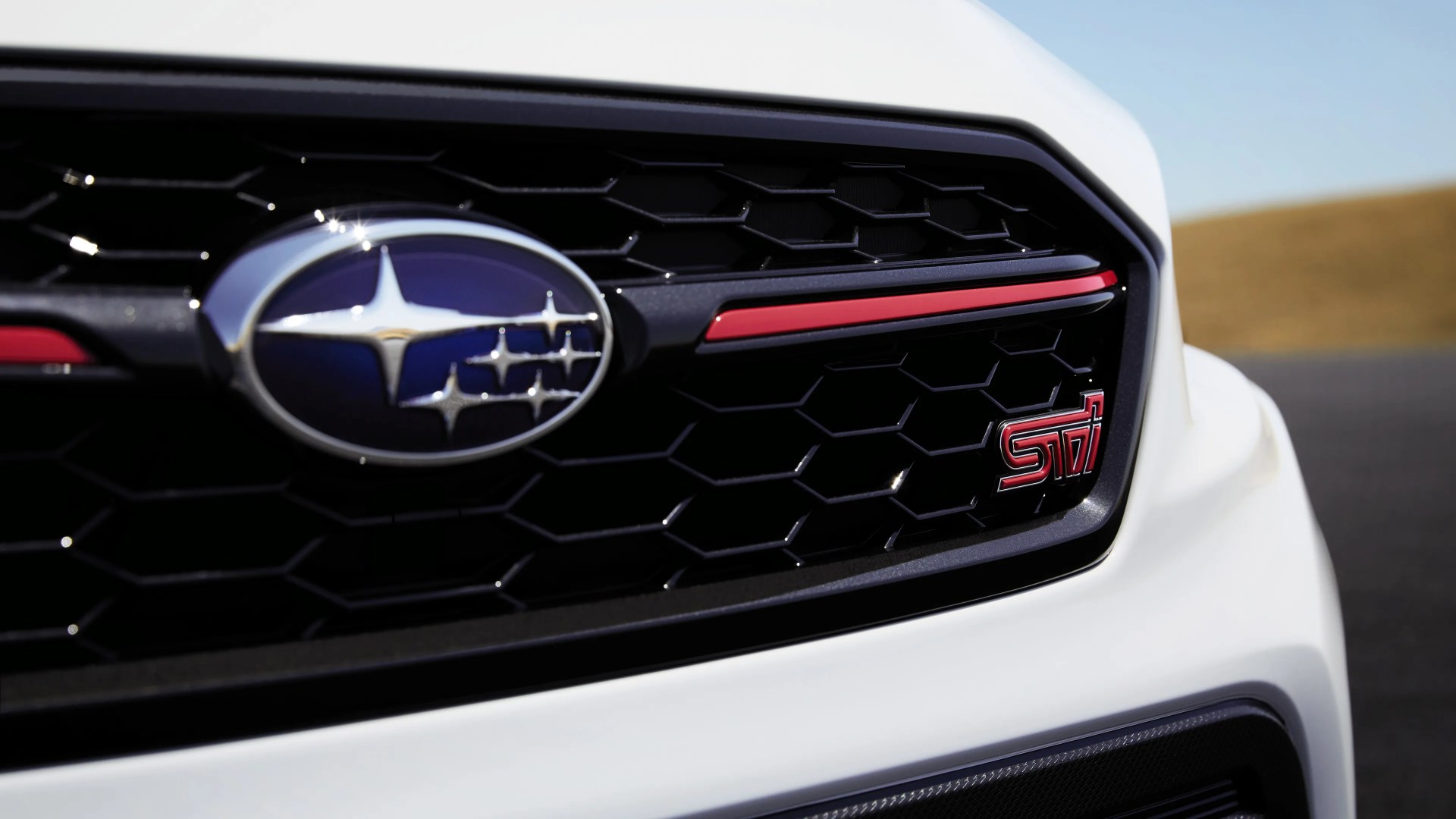 Close-up of a white Subaru car front grille featuring the Subaru emblem on the left and a red "STI" badge on the right, with a honeycomb pattern and a red accent line above the badge.