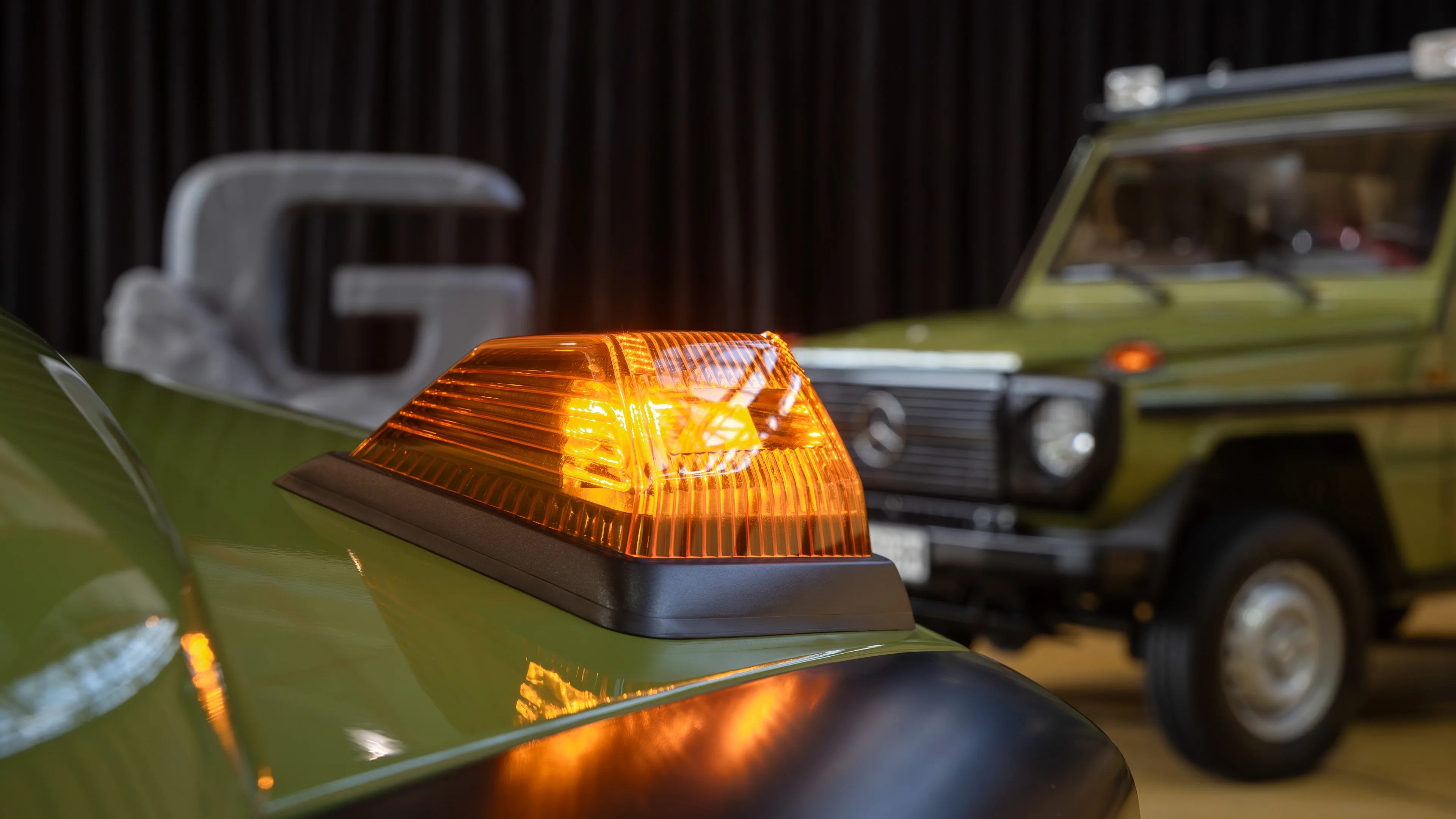 Close-up of an amber-colored emergency or warning light mounted on the hood of a green vehicle, with another green vehicle and a large blurred "G" sculpture in the background.