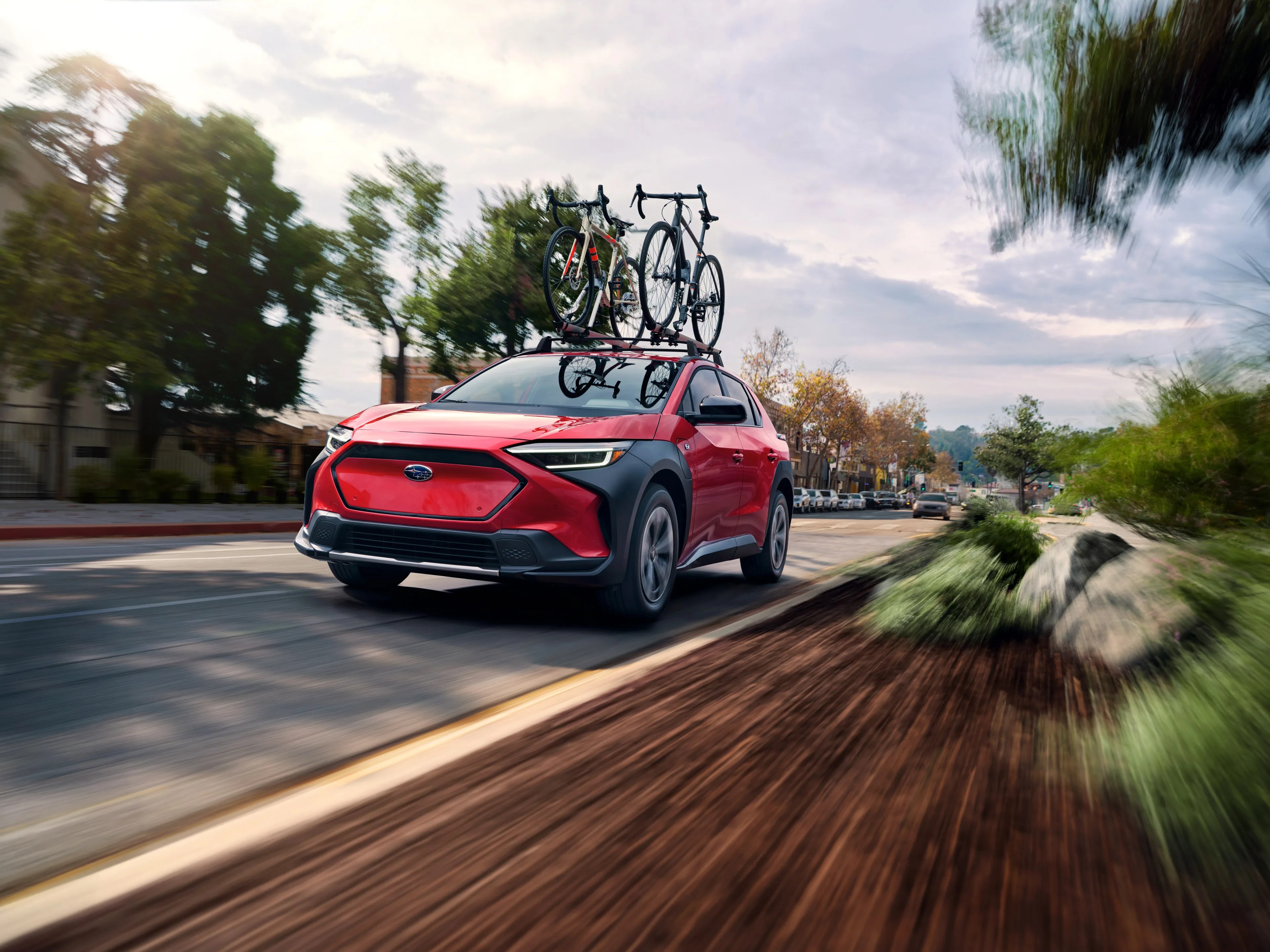 Red Subaru SUV driving on a street with two bicycles mounted on the roof rack, surrounded by trees and buildings under a cloudy sky.