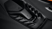 Close-up of a black McLaren car's aerodynamic engine cover featuring multiple air vents and a transparent section revealing part of the engine, with a carbon fiber textured surface surrounding it.
