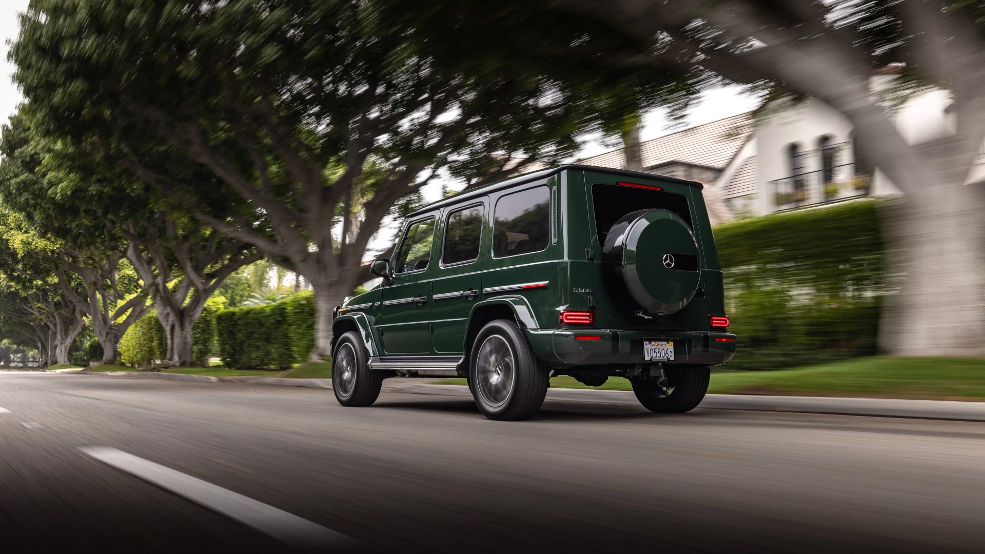 Dark green Mercedes-Benz G-Class SUV driving on a tree-lined suburban street, viewed from the rear right side. The vehicle has a spare tire mounted on the back door and tinted windows. The background shows large trees and residential houses with hedges.
