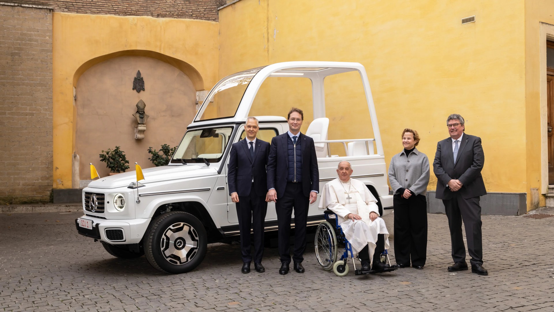 A white Mercedes-Benz popemobile with a transparent roof and two small yellow flags on the front is parked on a cobblestone surface. Five people stand in front of the vehicle, including a man in white papal robes seated in a wheelchair. The background features yellow and beige walls with an arched niche containing a bust and some greenery.