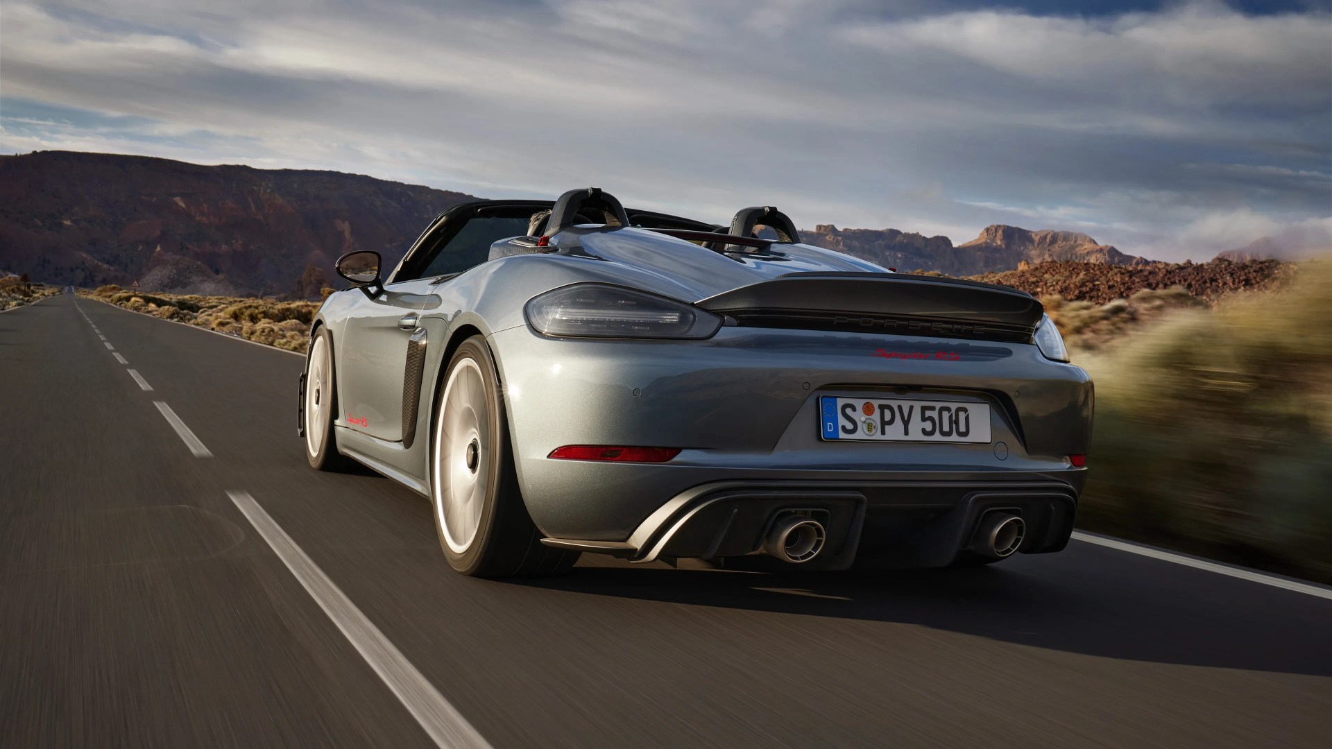Silver Porsche Spyder RS convertible sports car driving on an open road with mountainous terrain in the background under a partly cloudy sky. The car has a rear spoiler, dual exhaust pipes, and a German license plate.