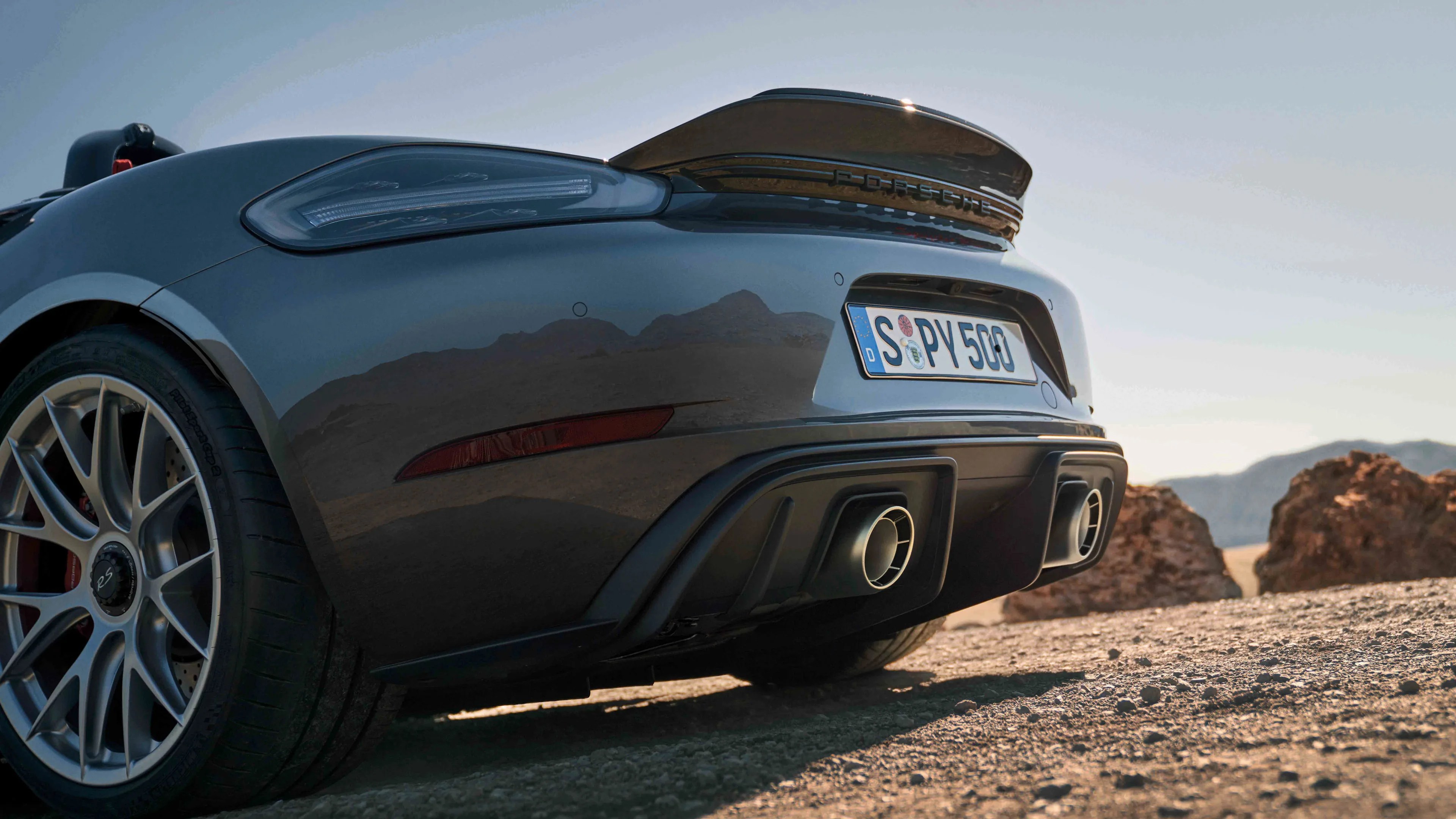 Rear view of a dark gray Porsche sports car with a raised rear spoiler, dual exhaust pipes, and a visible "RS" emblem on the alloy wheel, parked on a gravel surface with rocky terrain and mountains in the background.