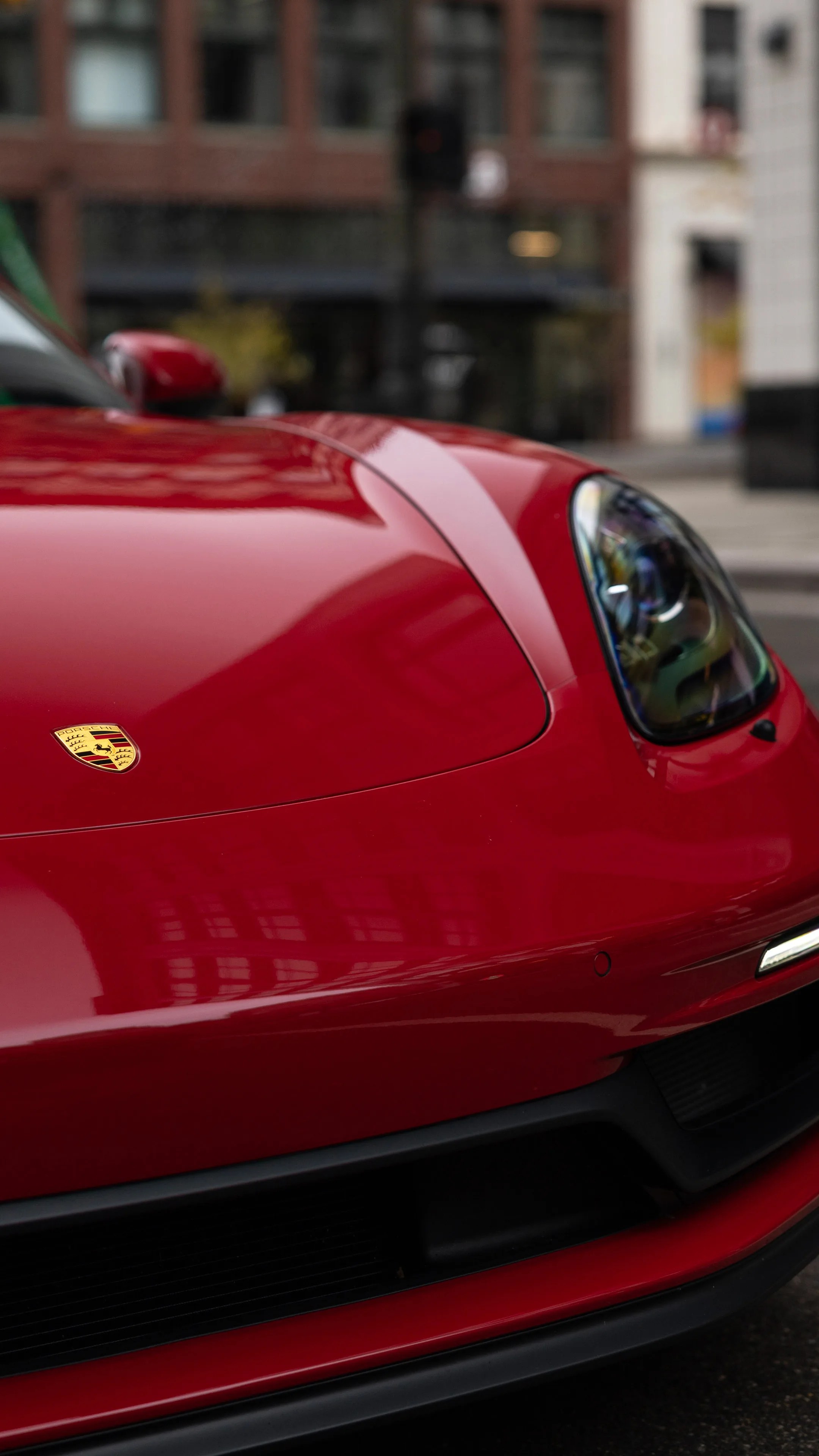 Close-up of the front end of a red Porsche sports car, showing the hood with the Porsche emblem, the right headlight, and part of the front bumper and grille. The car's glossy surface reflects nearby buildings.