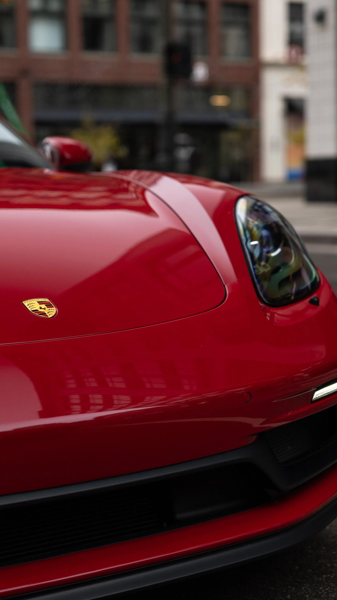 Close-up of the front end of a red Porsche sports car, showing the hood with the Porsche emblem, the right headlight, and part of the front bumper and grille. The car's glossy surface reflects nearby buildings.