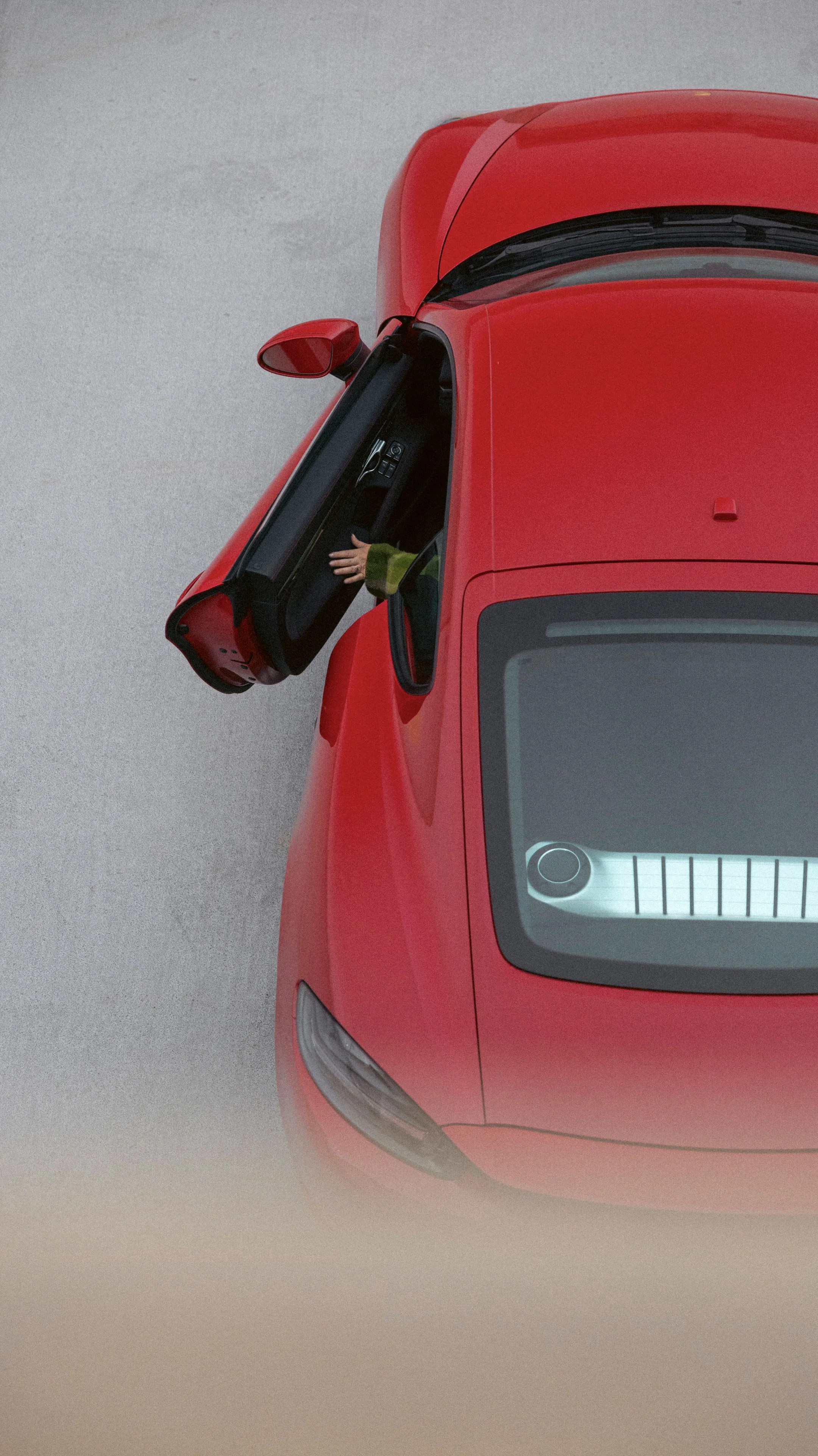 Top-down view of a red sports car with the driver's side door open. A person's hand, wearing a green sleeve, is visible reaching out from inside the car. The car is parked on a light gray surface.