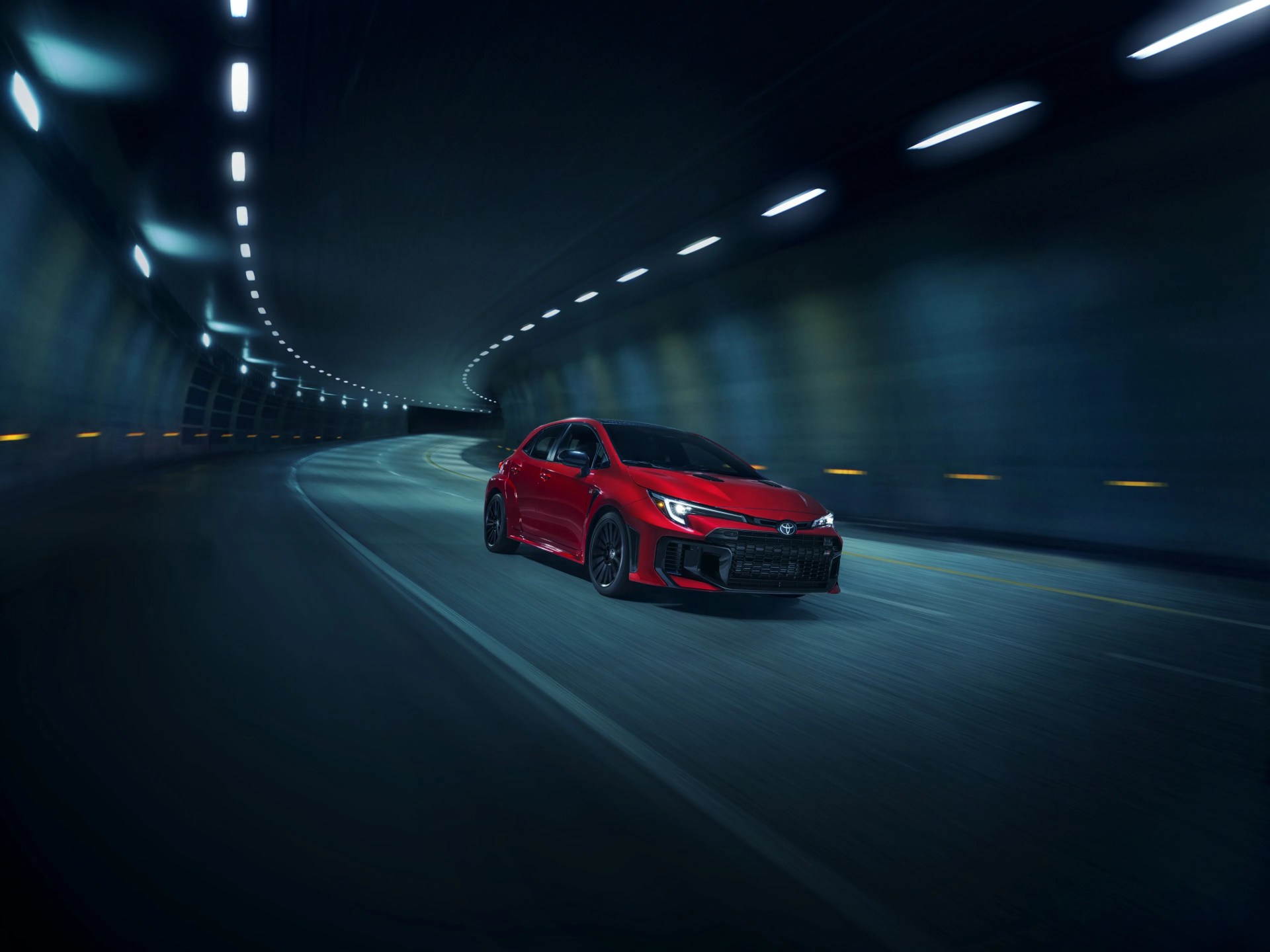 Red sporty sedan driving through a dimly lit tunnel with curved walls and overhead lights, creating a sense of motion and speed.